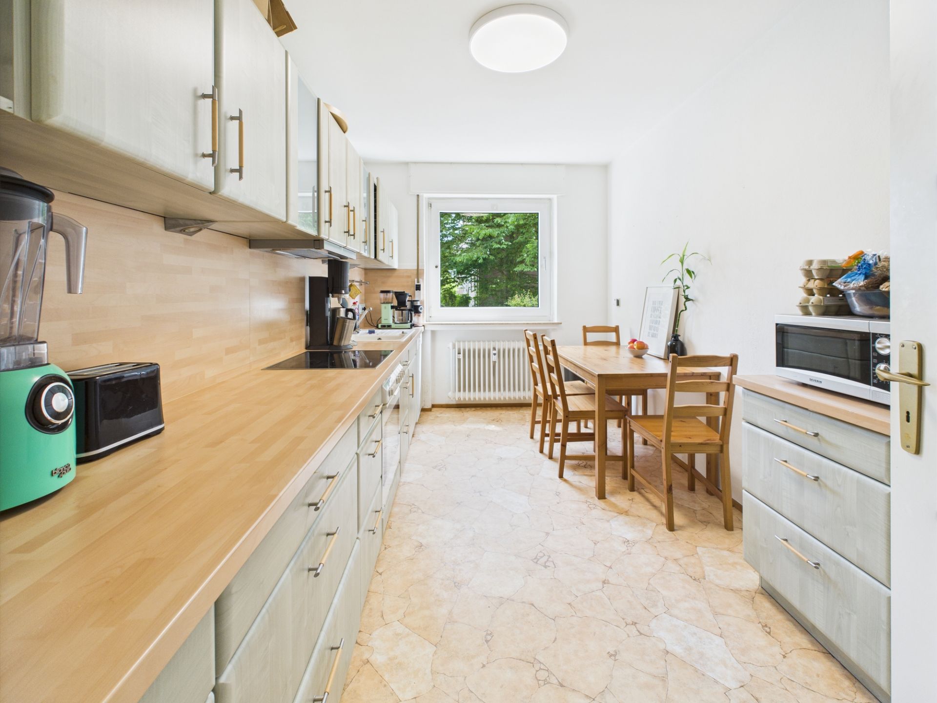 Helle Küche mit Holzmöbeln, Geräten, Fenster und Esstisch mit vier Stühlen.