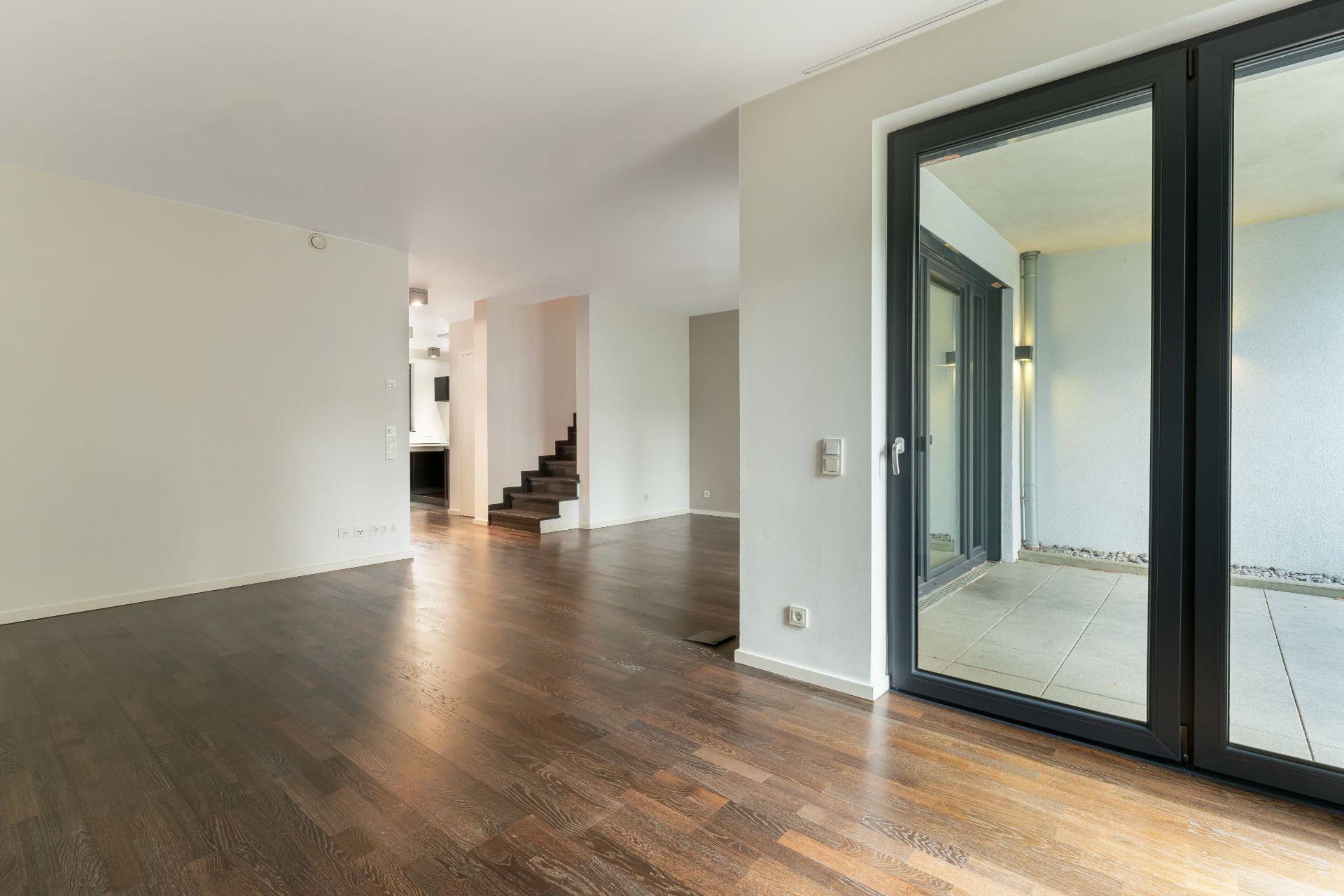 Leeres Wohnzimmer mit Holzboden, Glastür und offener Treppe im Hintergrund.