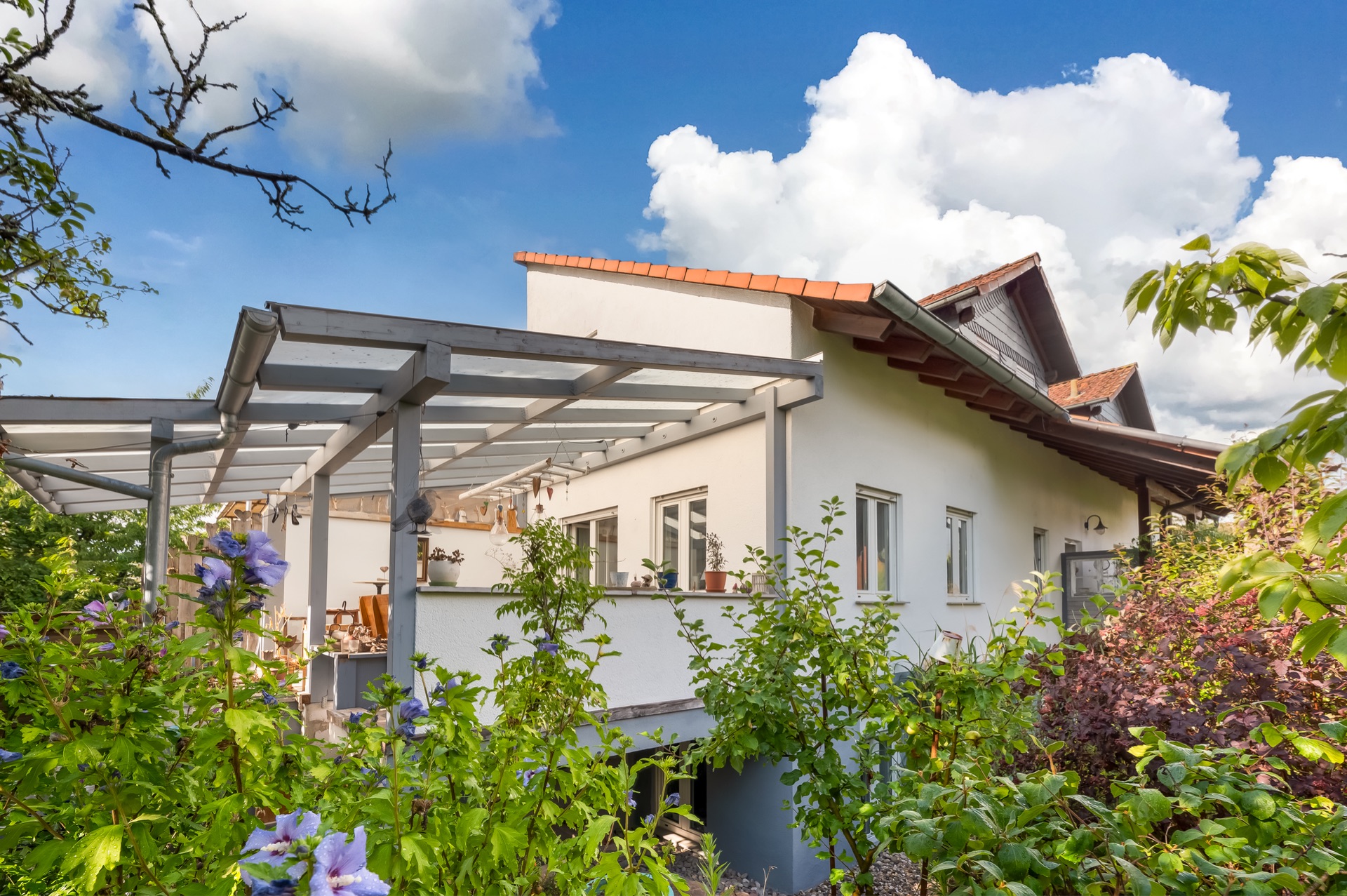 Helles Haus mit Terrasse, Glasdach und blühenden Sträuchern im Garten, unter blauem Himmel.