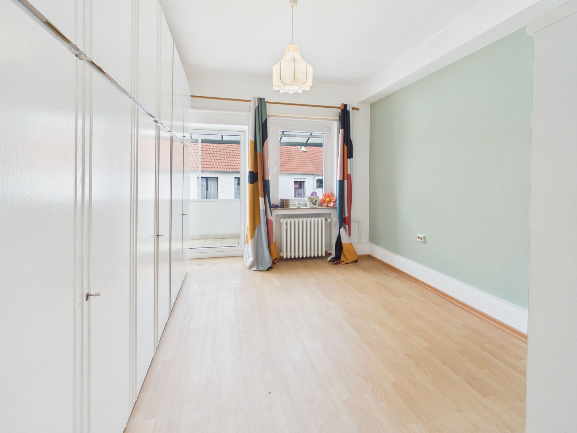 Helles Zimmer mit Holzfußboden, Einbauschränken, Fenster und farbigen Vorhängen.