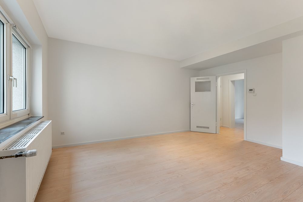 Helles, leeres Zimmer mit Holzfußboden, Fenster und Heizkörper auf der linken Seite.
