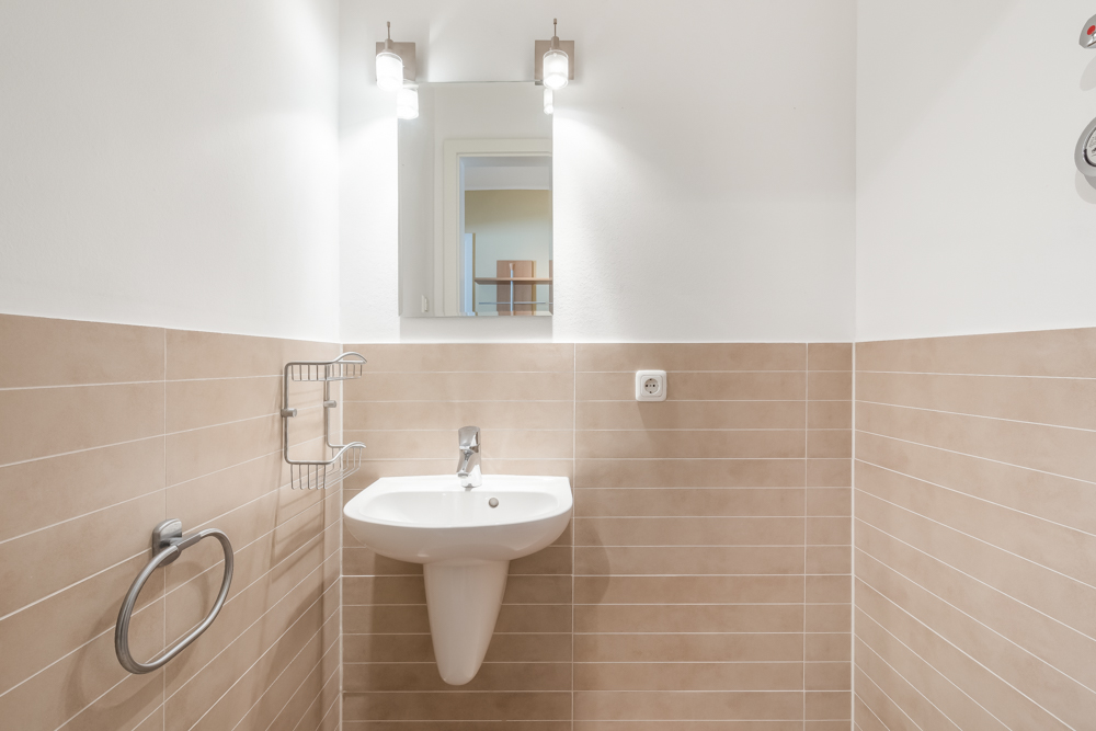 Helles Badezimmer mit Waschbecken, Spiegel, zwei Lampen und beigen Wandfliesen.