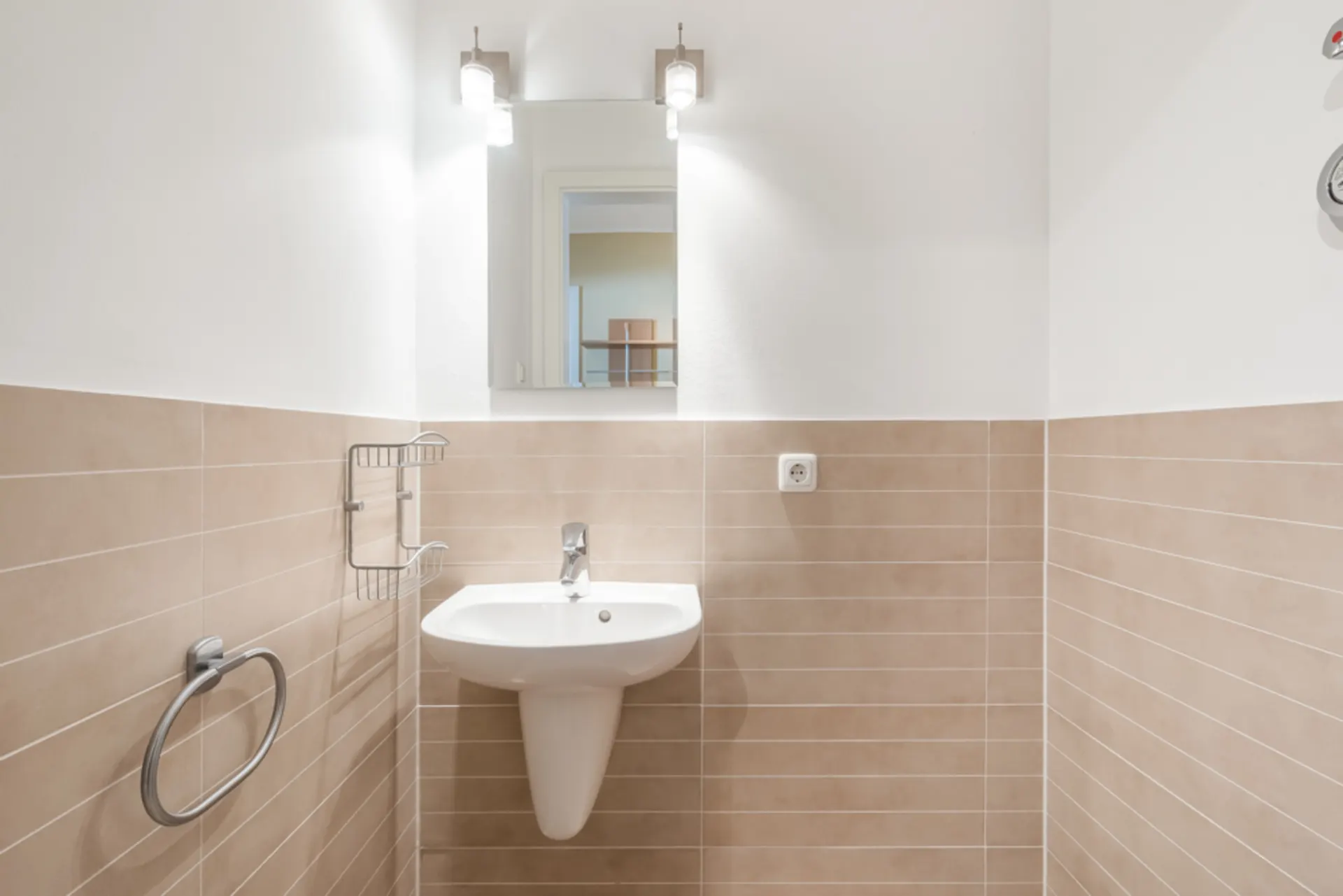 Helles Badezimmer mit Waschbecken, Spiegel, zwei Lampen und beigen Wandfliesen.