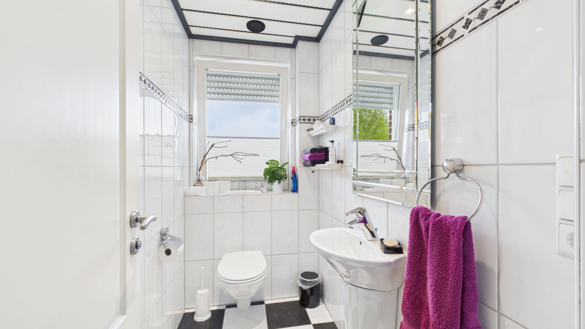 Helles Badezimmer mit WC, Waschbecken, Spiegel, Fenster und lila Handtuch.