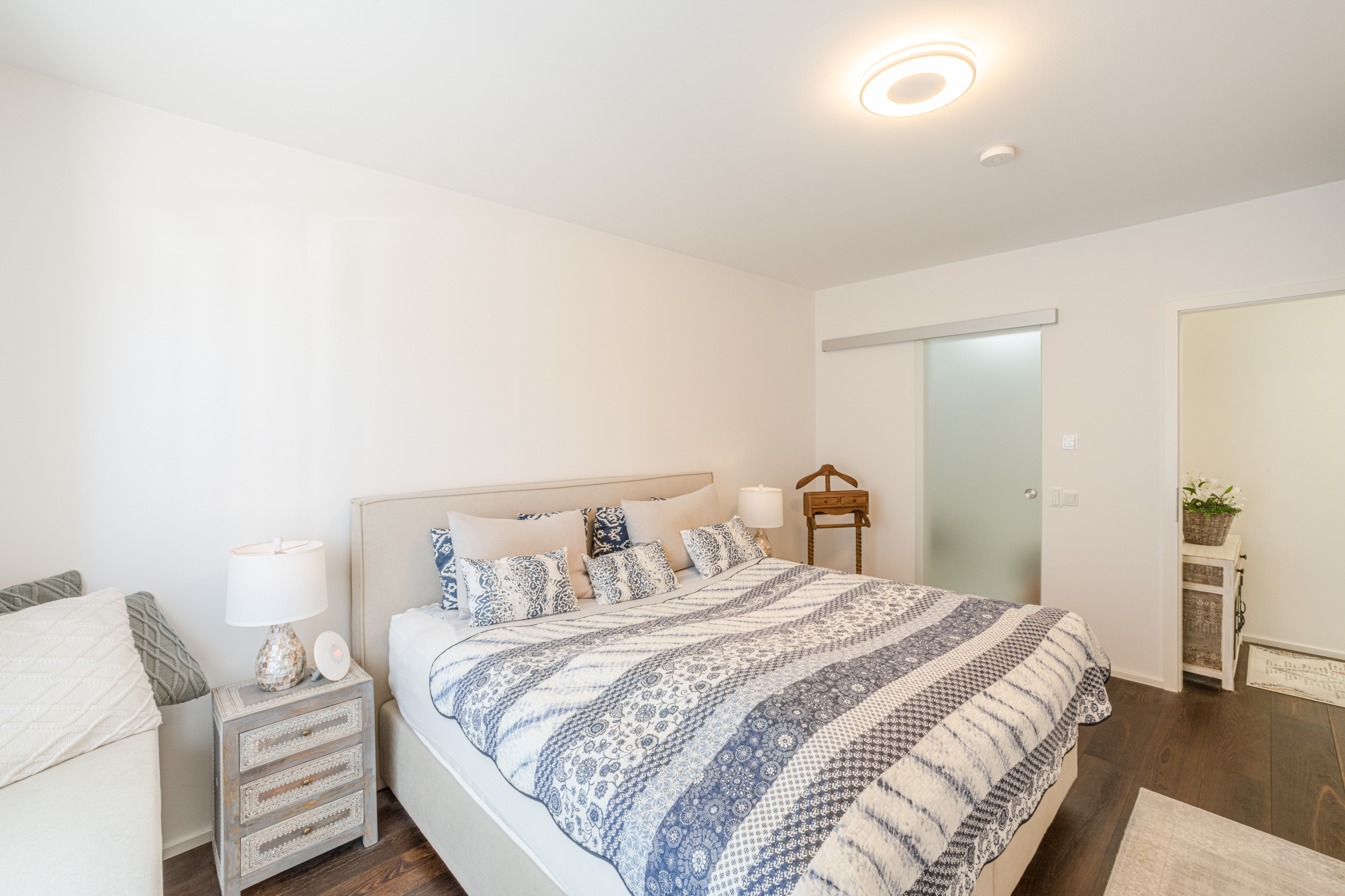 Helles Schlafzimmer mit Doppelbett, Nachttisch, Stehlampe und gemustertem Bettbezug.
