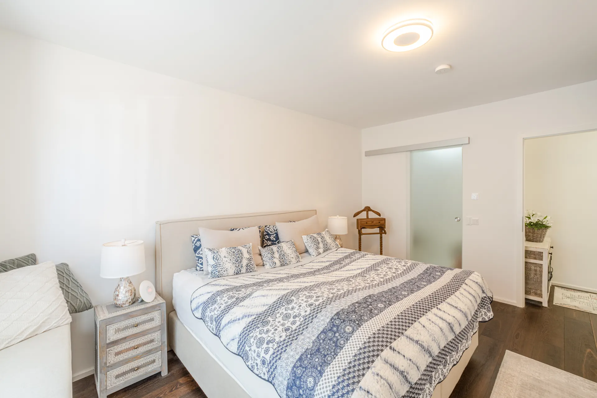 Helles Schlafzimmer mit Doppelbett, Nachttisch, Stehlampe und gemustertem Bettbezug.