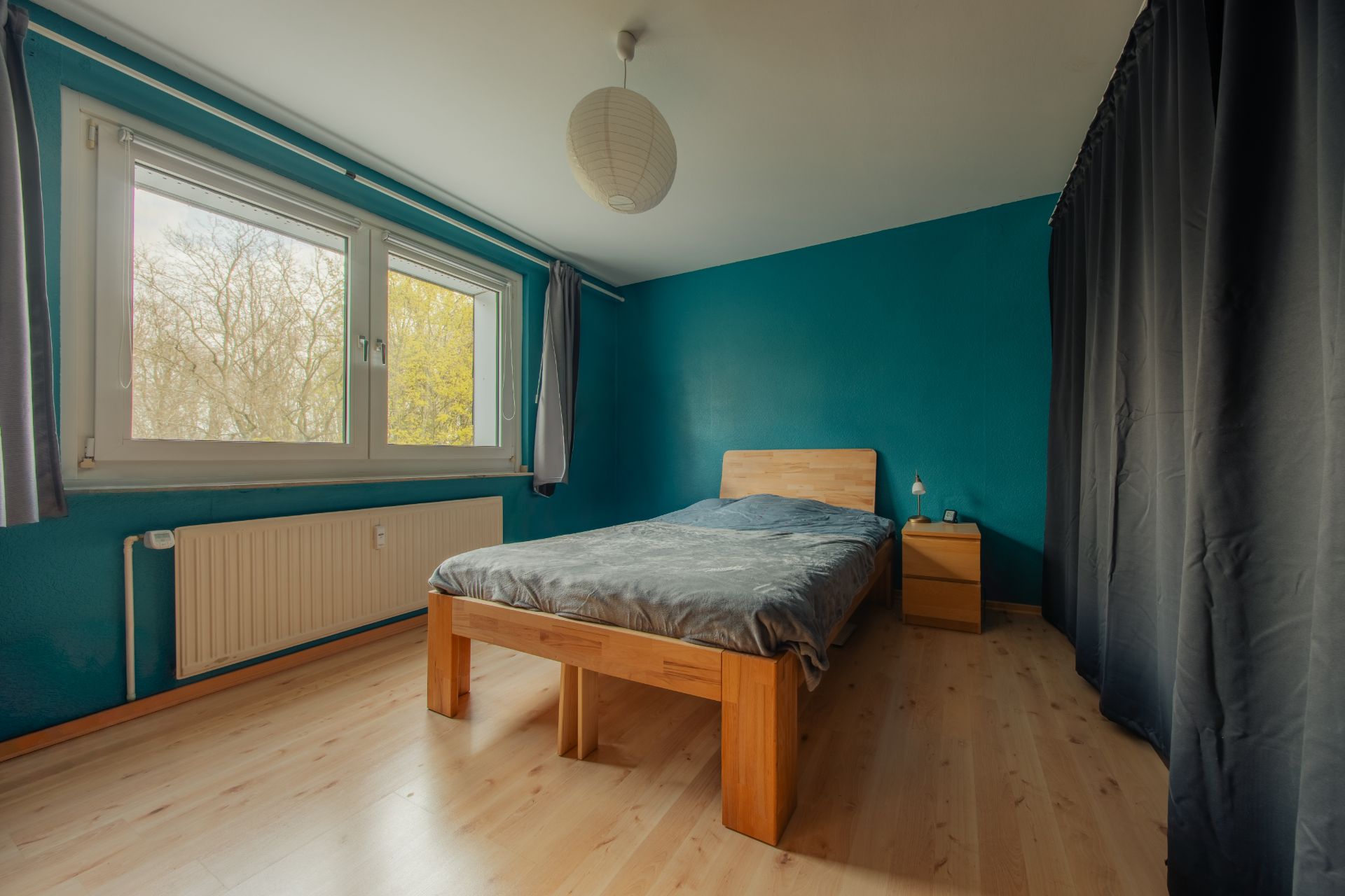 Schlafzimmer mit Holzbett, Nachttisch, großem Fenster und blauen Wänden.