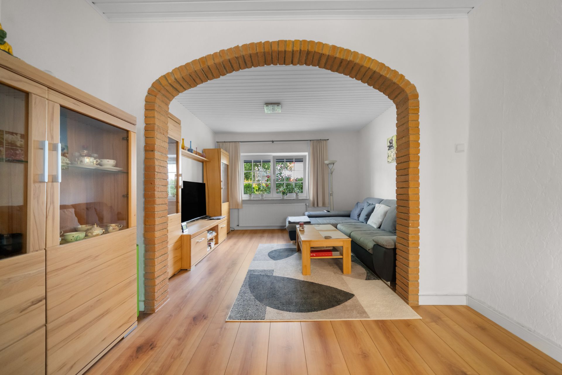Wohnzimmer mit Holzfußboden, Sofa, Couchtisch und Glastürenschrank, durch einen Backsteinbogen getrennt.