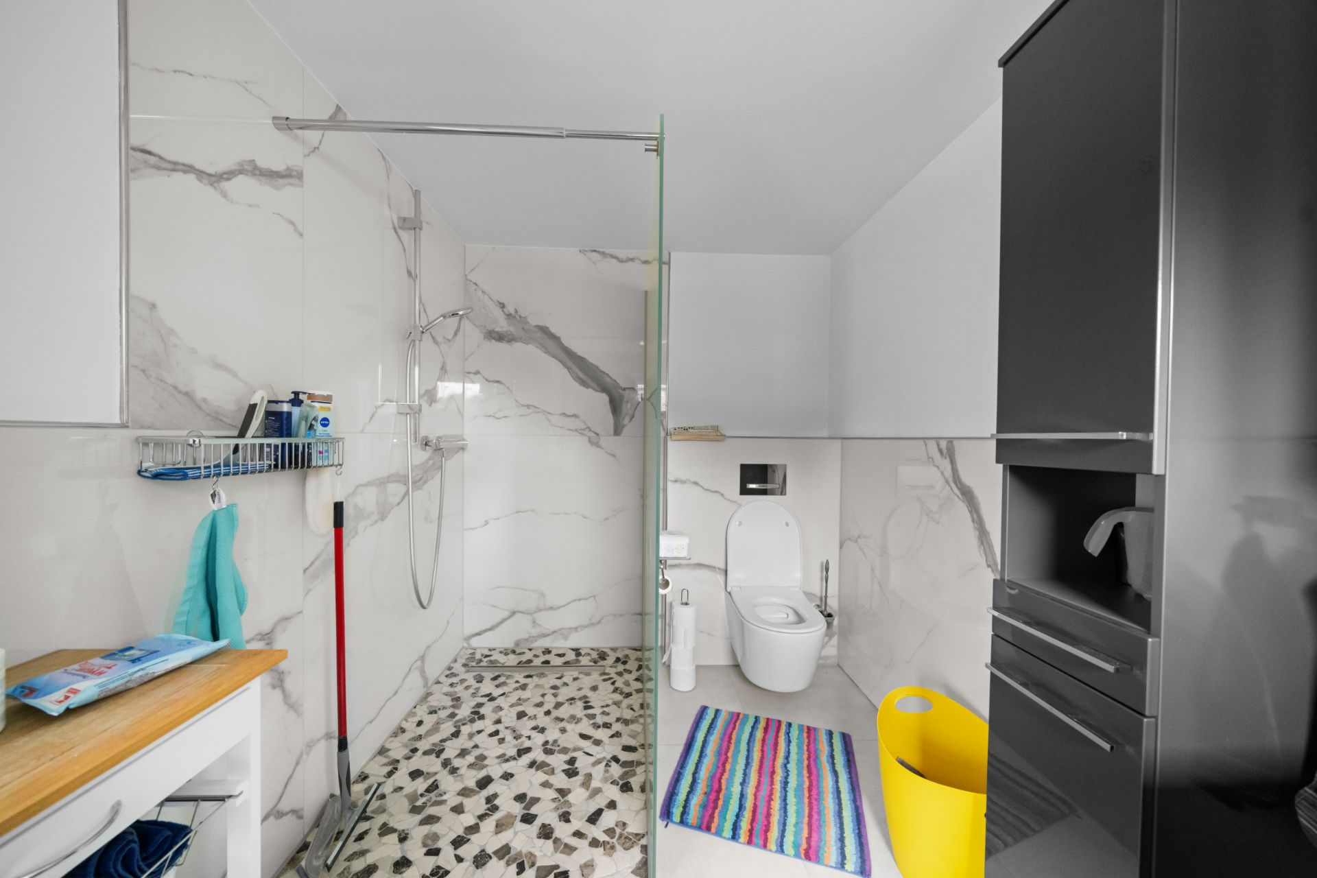 Modernes Badezimmer mit Dusche, WC, buntem Teppich und gelbem Wäschekorb.