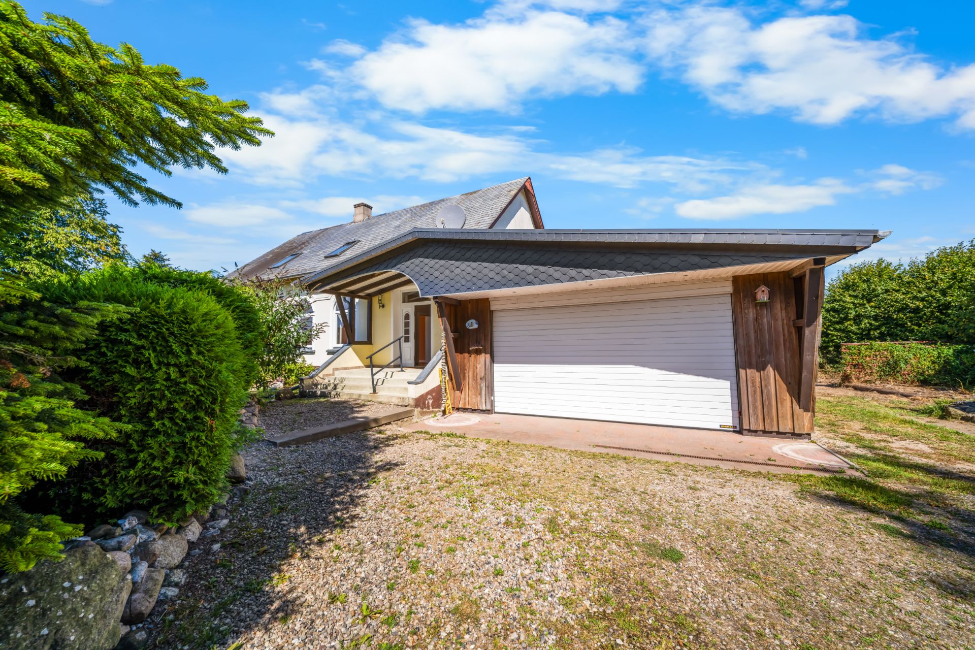 Einfamilienhaus mit schrägem Dach und Garage, umgeben von Bäumen und Garten.