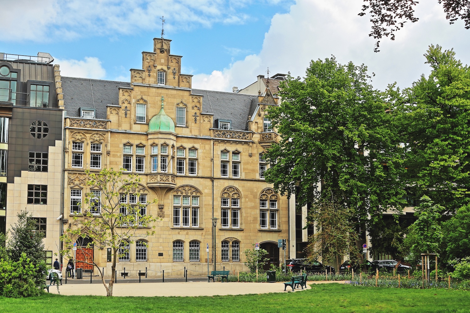Historisches Wohngebäude mit Verzierung, davor Bäume und eine Parkbank auf einer Grünfläche.