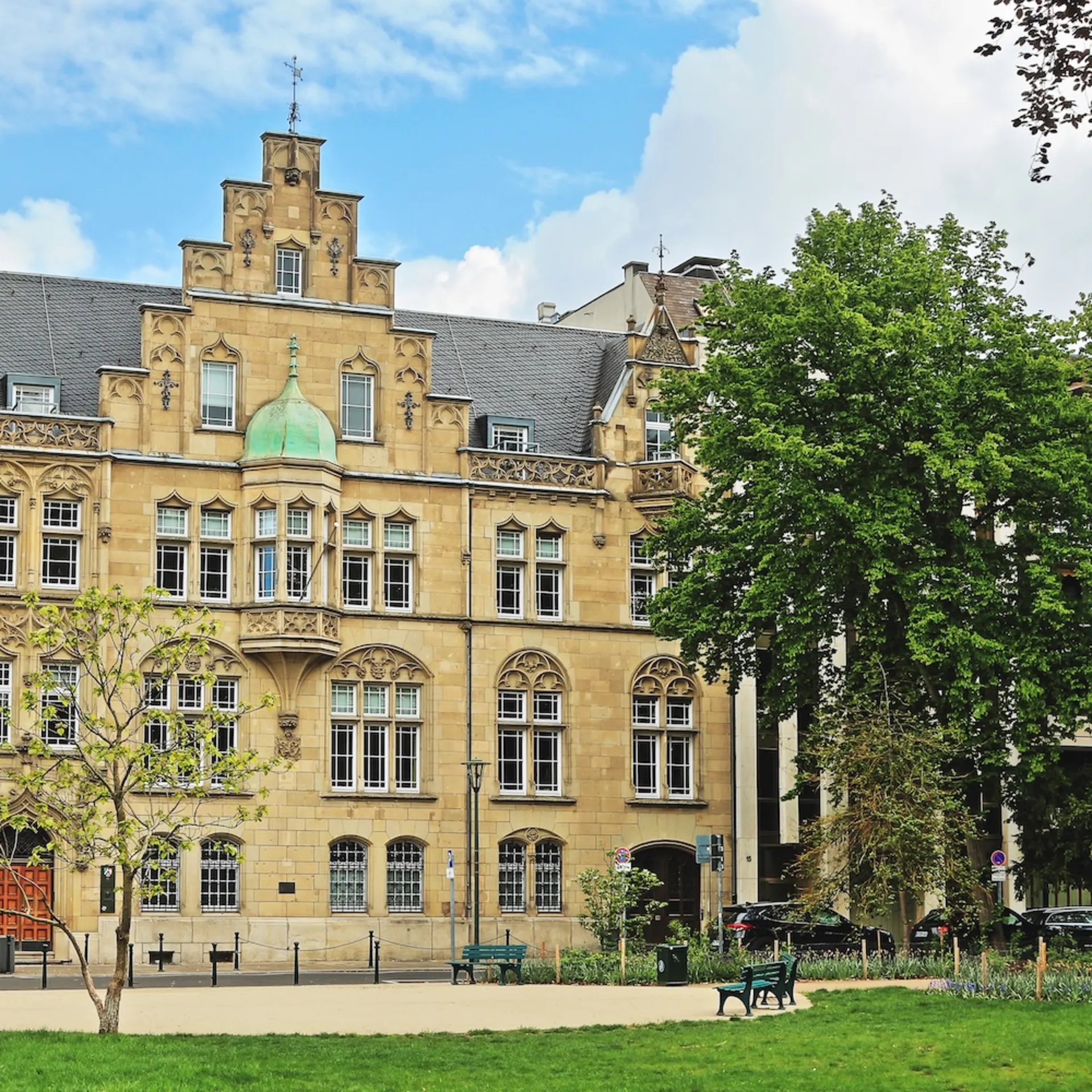 Historisches Wohngebäude mit Verzierung, davor Bäume und eine Parkbank auf einer Grünfläche.