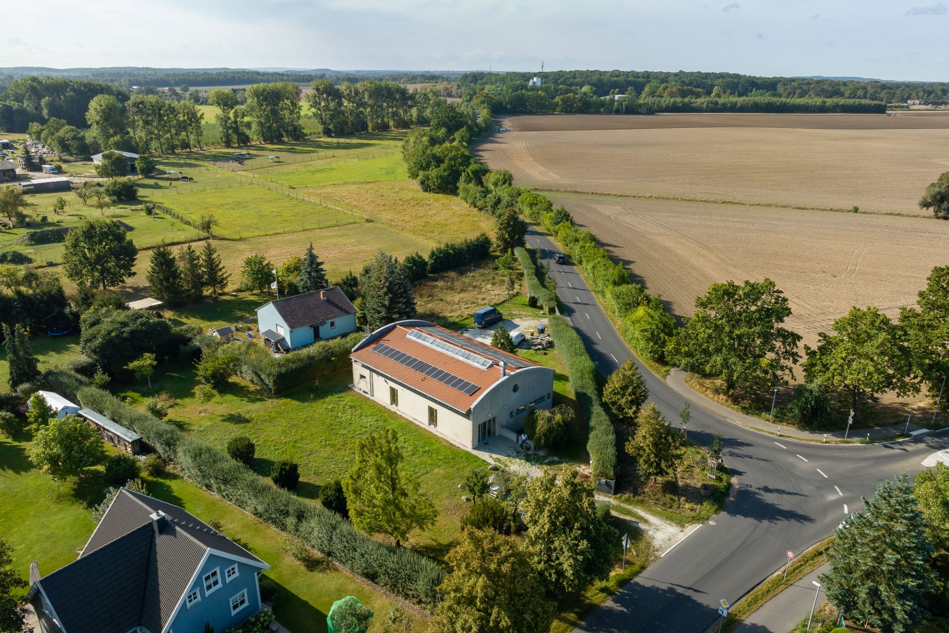 Luftaufnahme von Wohnhäusern mit Gärten an einer Landstraße, angrenzend an Felder und Bäume.
