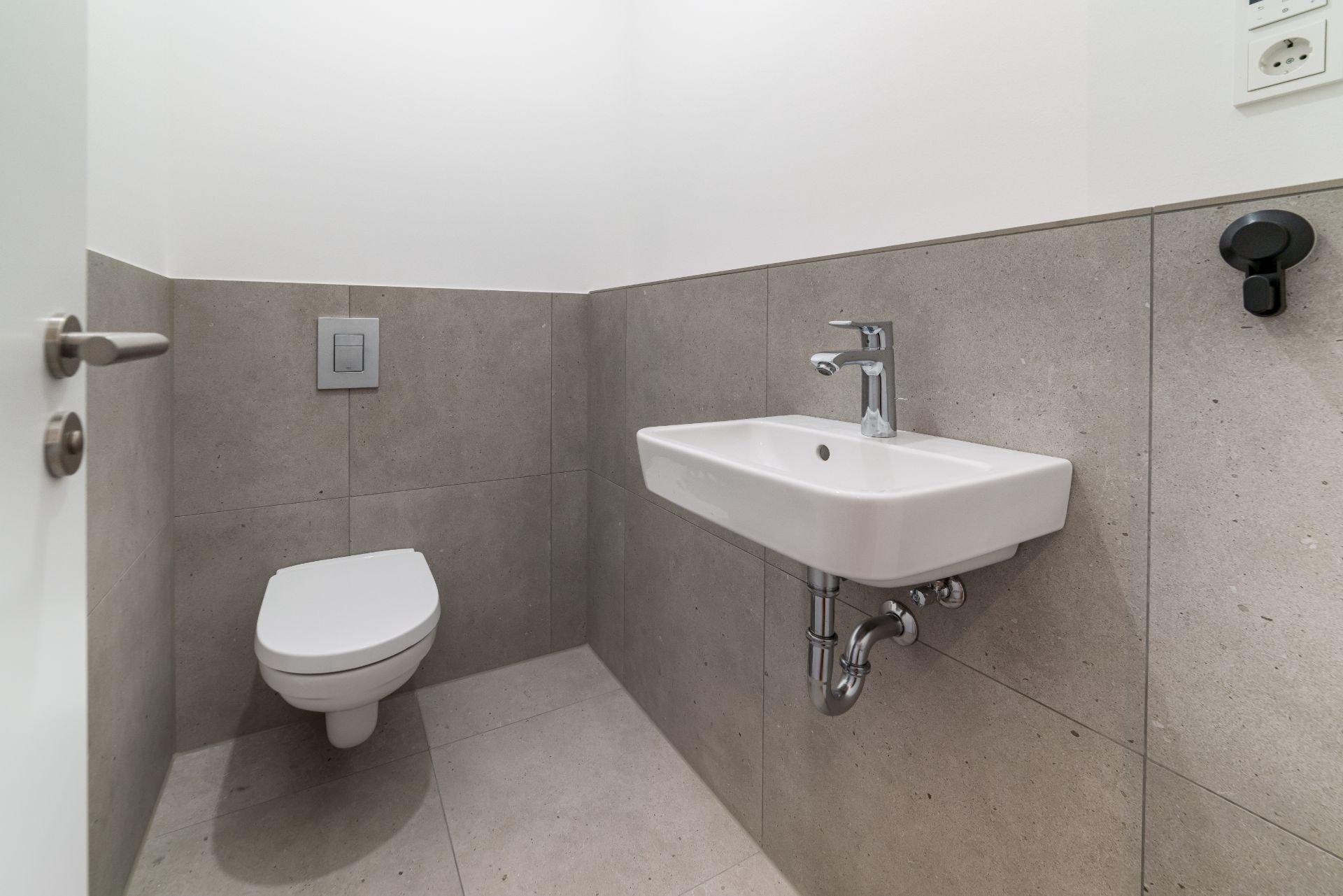 Modernes Badezimmer mit wandhängender Toilette und Waschbecken, graue Fliesen an Wand und Boden.