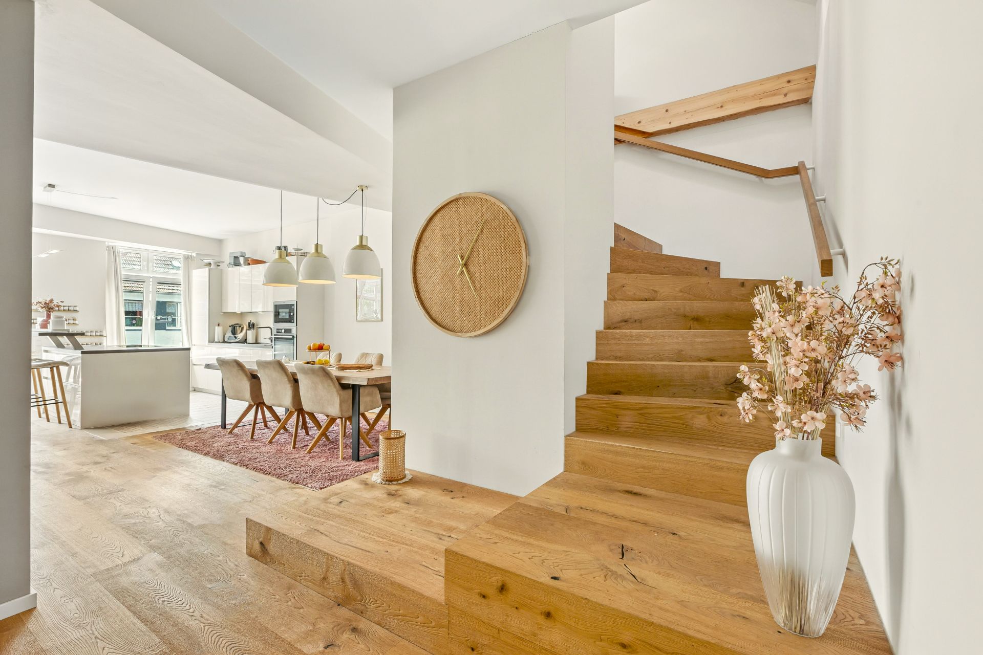 Helles Wohnzimmer mit Holztreppe, Esstisch, offener Küche und Vase mit Blumen auf dem Boden.