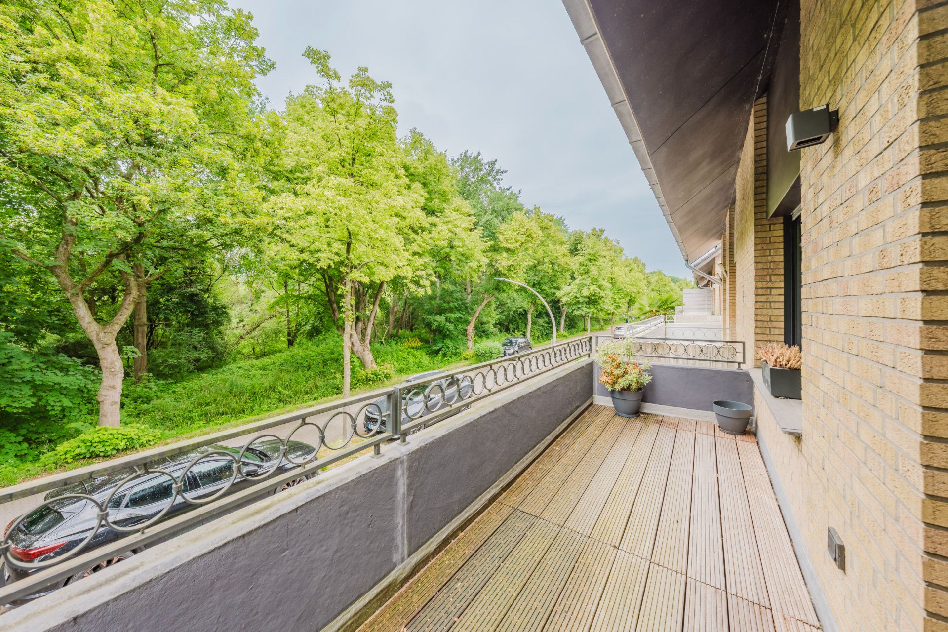 Großer Balkon mit Holzboden, Blick auf grüne Bäume und parkende Autos an einer Straße.