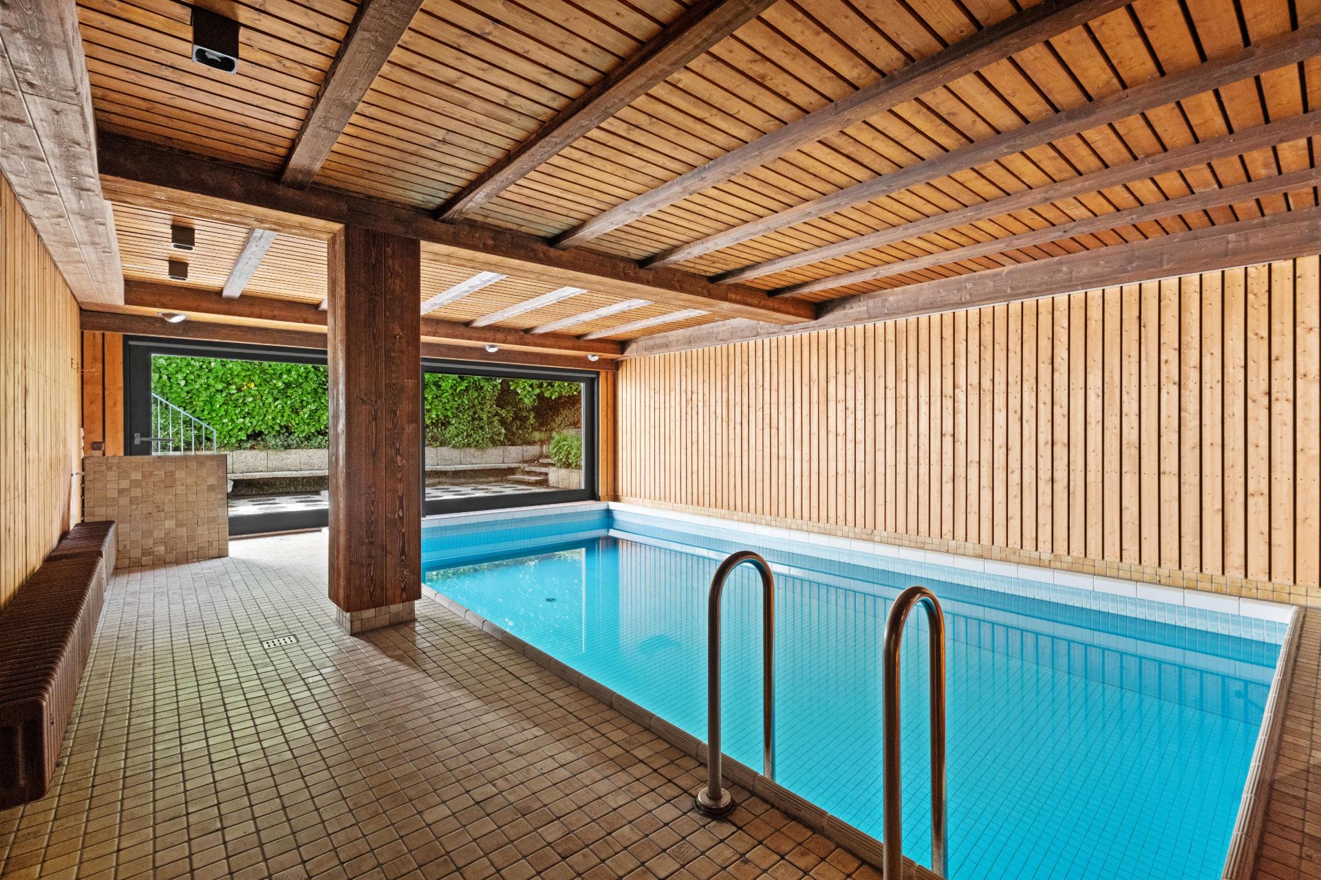 Überdachtes Schwimmbecken mit Holzdecke und -wänden, großes Fenster mit Blick ins Grüne.