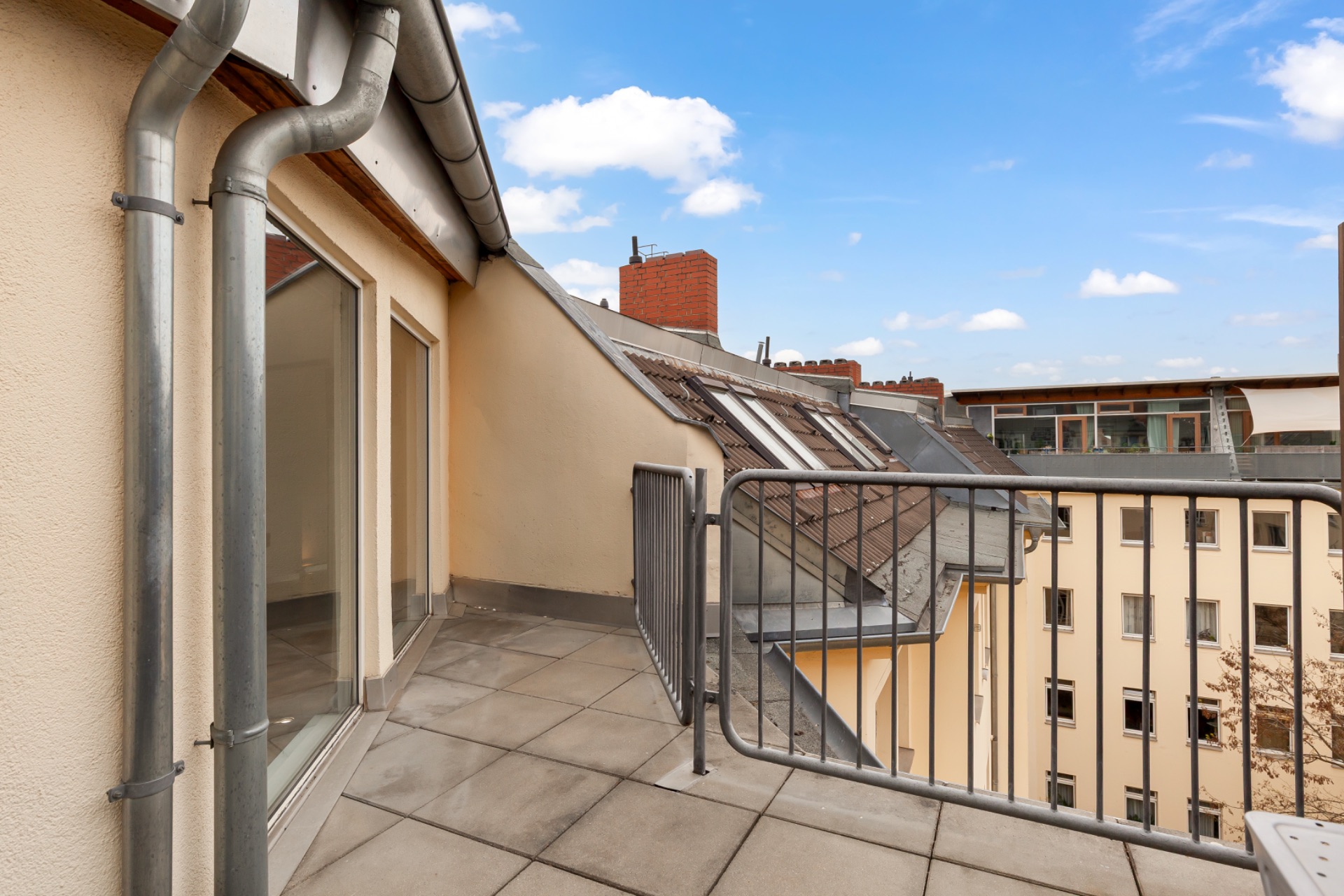 Kleine Dachterrasse mit Geländer und Blick auf Nachbargebäude unter blauem Himmel.