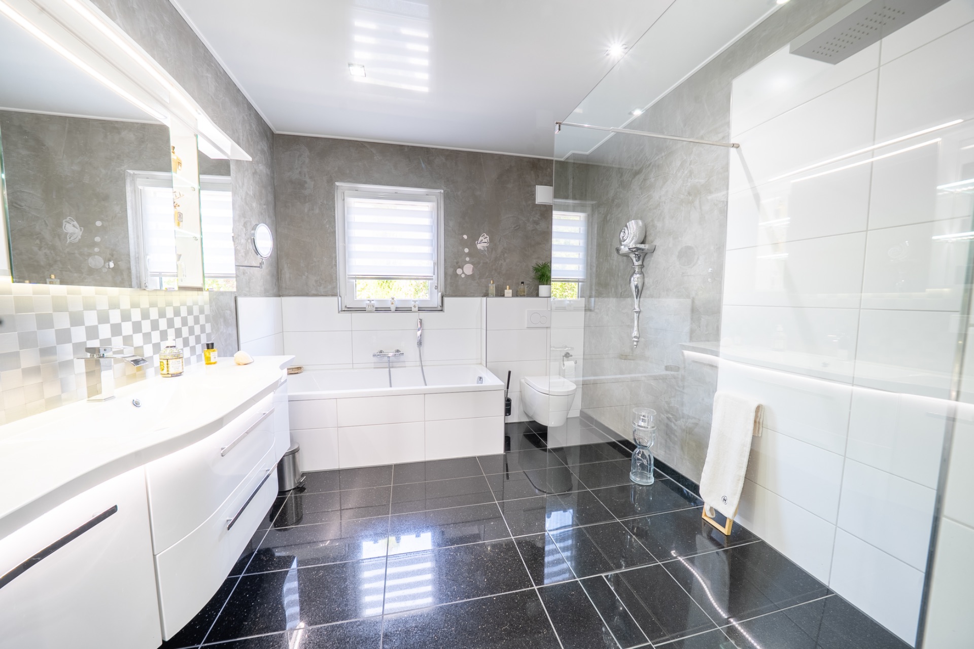 Modernes Badezimmer mit Badewanne, Dusche, Waschbecken und Fenster, weiße Fliesen und schwarze Bodenfliesen.