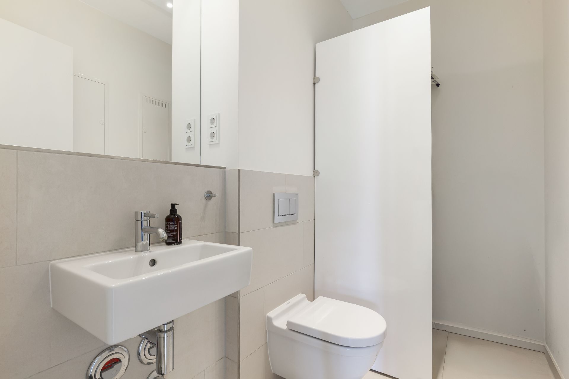 Modernes Badezimmer mit Wandwaschbecken, Spiegel und Toilette, helle Fliesen und weiße Tür.