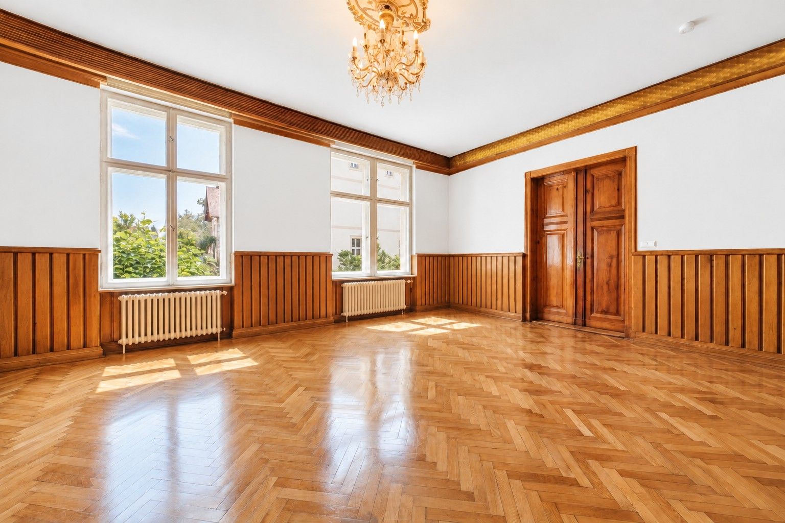 Geräumiges, leeres Zimmer mit Parkett, großen Fenstern, Kronleuchter und Holztüren.