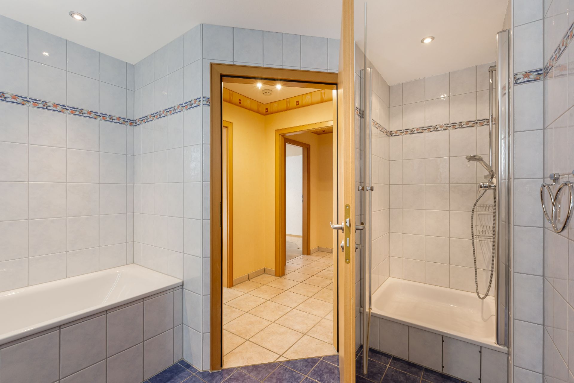 Badezimmer mit Badewanne und Dusche, Tür führt zu einem Flur mit Fliesenboden.