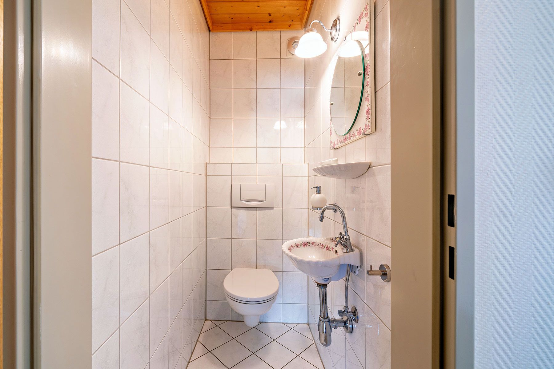 Kleines Bad mit Wandtoilette, Waschbecken, Spiegel und weiß gefliesten Wänden.