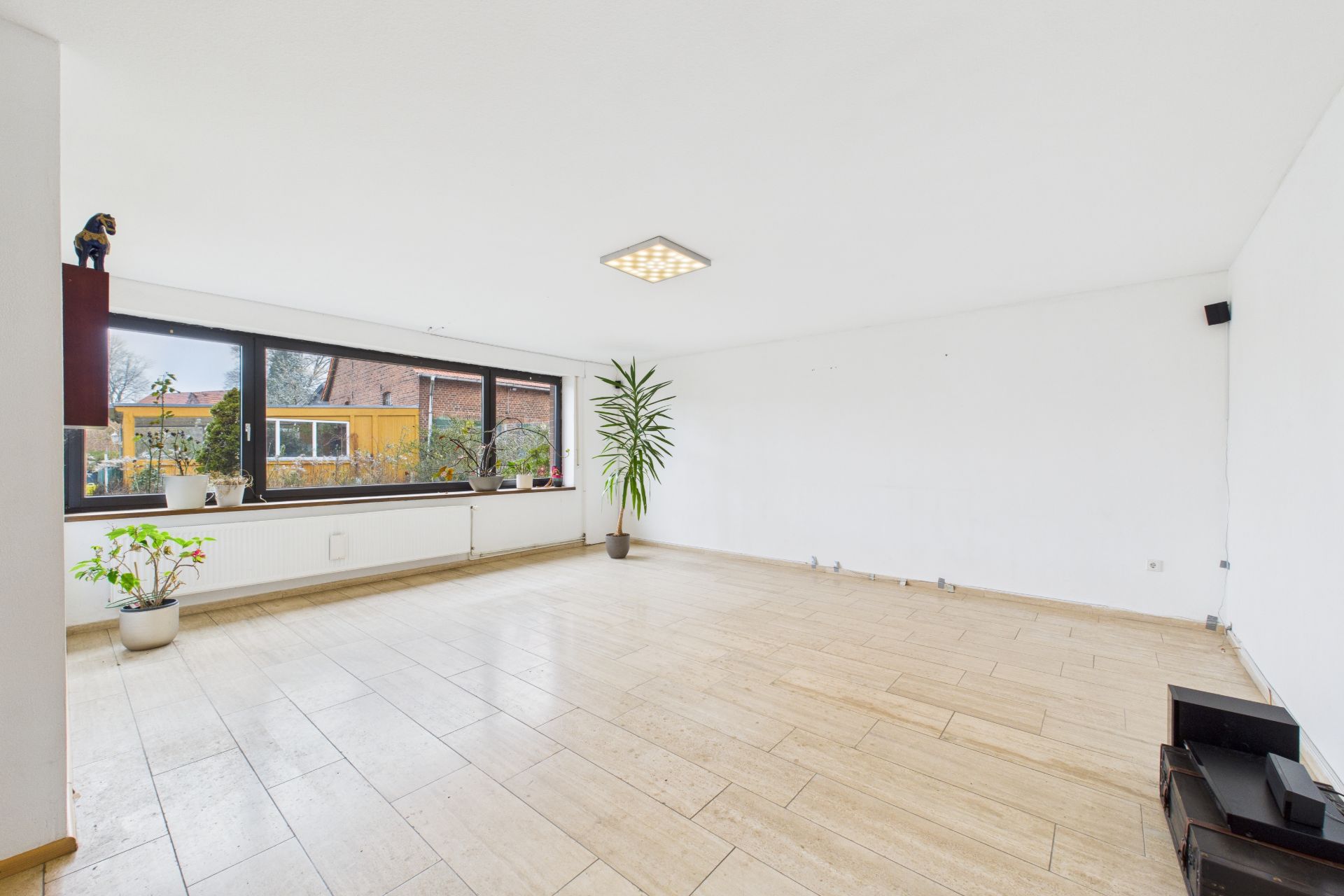 Helles, leerstehendes Wohnzimmer mit Fliesenboden, großen Fenstern und einigen Zimmerpflanzen.