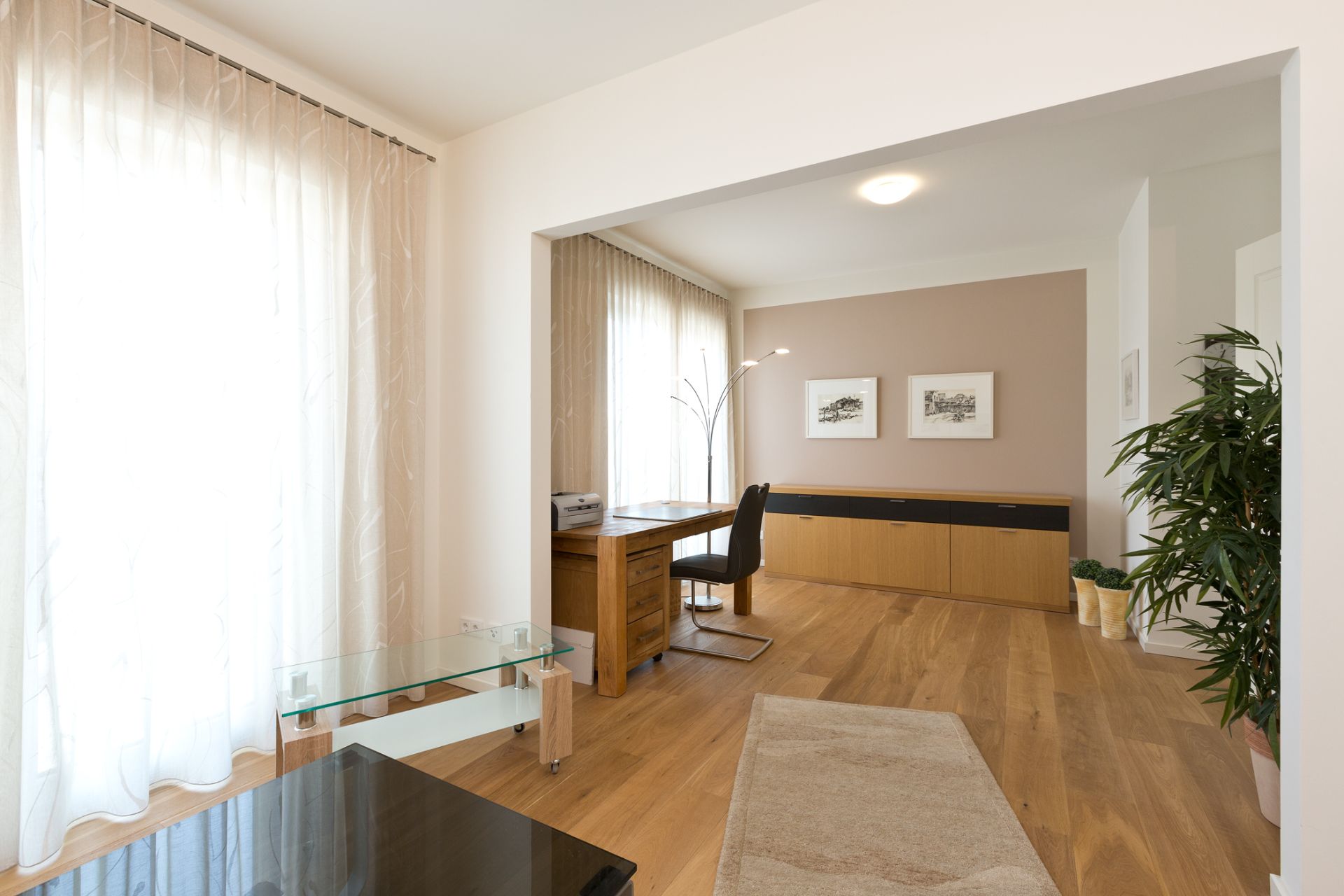 Helles Wohnzimmer mit Schreibtisch, Holzfußboden, Pflanzen und großen Fenstern mit Vorhängen.