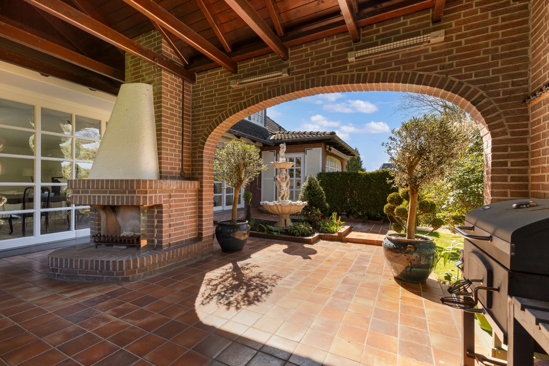 Überdachte Terrasse mit Kamin, Grill, Topfpflanzen und Blick auf einen Innenhof mit Springbrunnen.