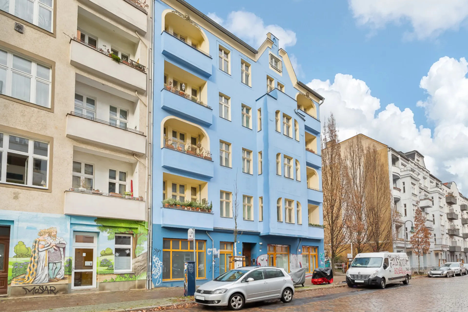 Mehrstöckiges Wohnhaus mit blauer Fassade und Balkonen an einer Straße mit geparkten Autos.