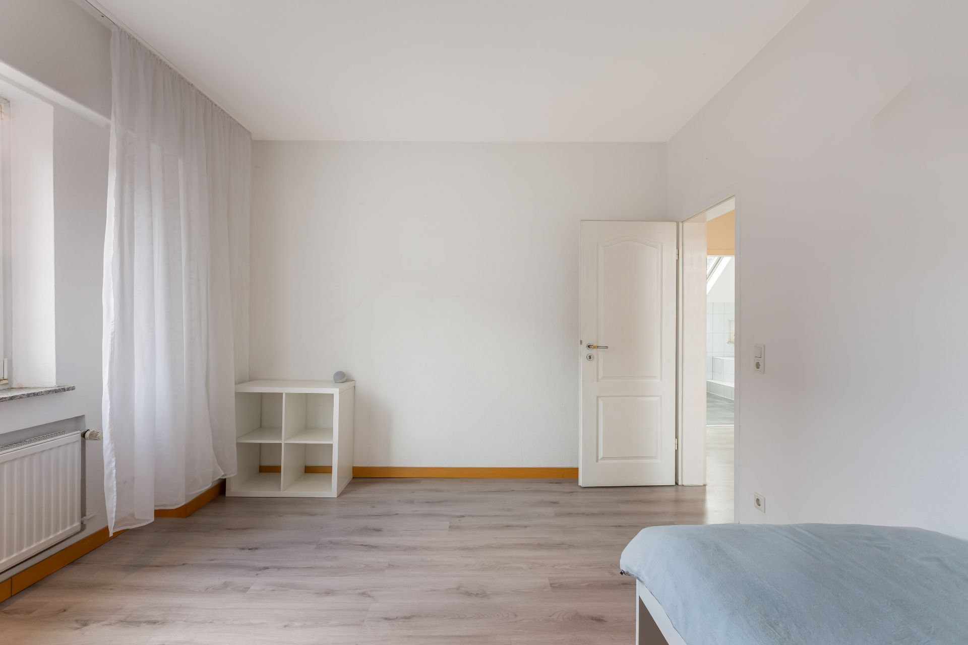 Helles Zimmer mit Holzfußboden, offenem Regal, Heizung und offen stehender Tür.