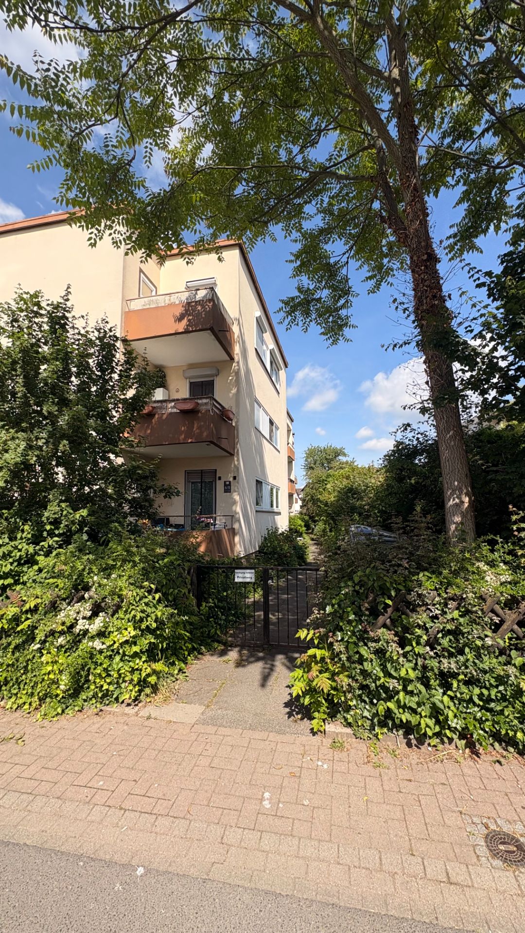 Dreistöckiges Wohnhaus mit Balkonen, Bäumen und Eingangstor im Vordergrund.