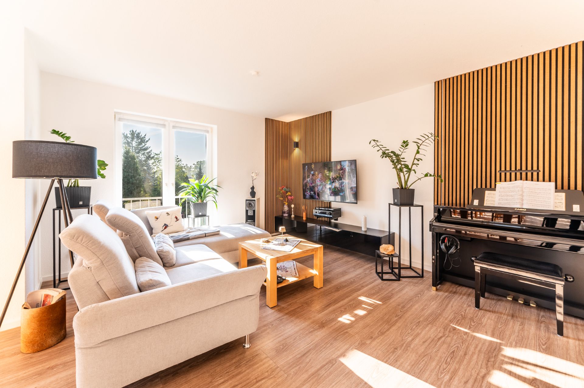 Helles Wohnzimmer mit Sofa, Couchtisch, TV, Stehlampe, Pflanzen und Klavier auf Holzfußboden.