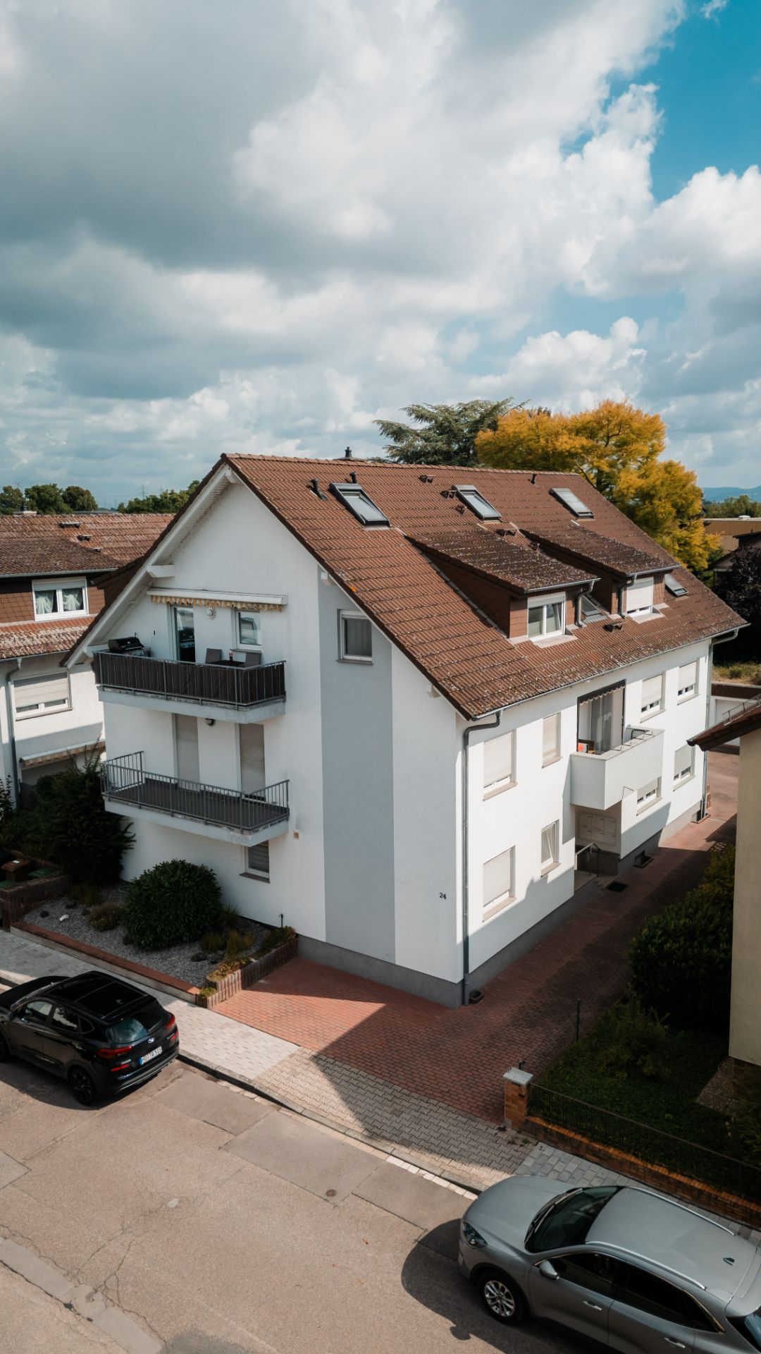 Modernes weißes Wohnhaus mit rotem Dach, mehreren Balkonen und geparkten Autos davor.