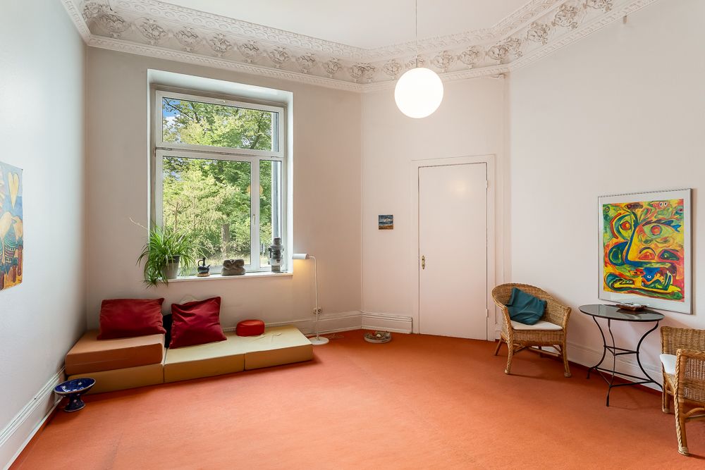 Wohnzimmer mit rotem Teppich, Sitzkissen, Fenster, Pflanzen und bunten Bildern an den Wänden.