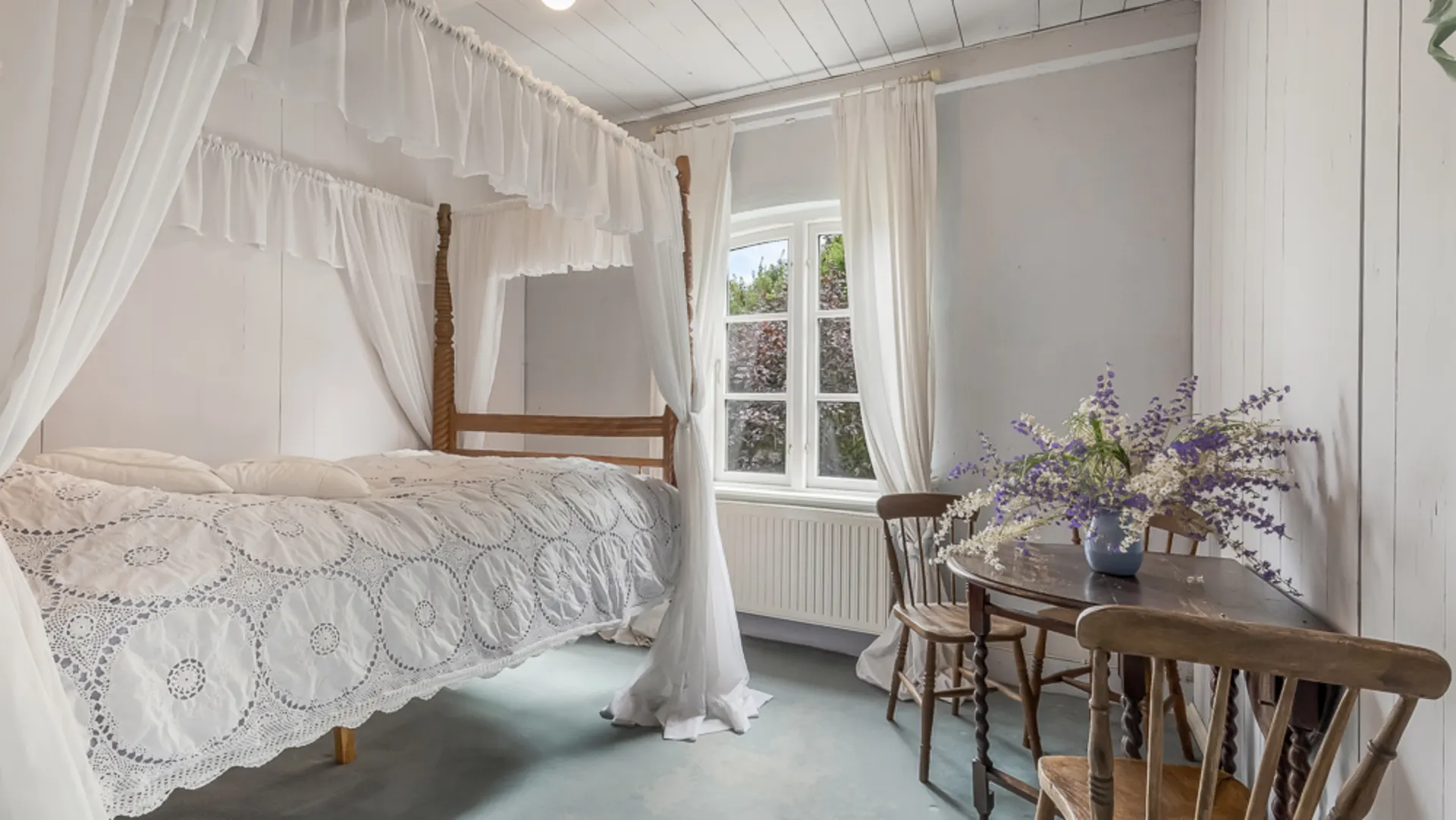 Helles Schlafzimmer mit Himmelbett, Holztisch, drei Stühlen und Blumenstrauß am Fenster.