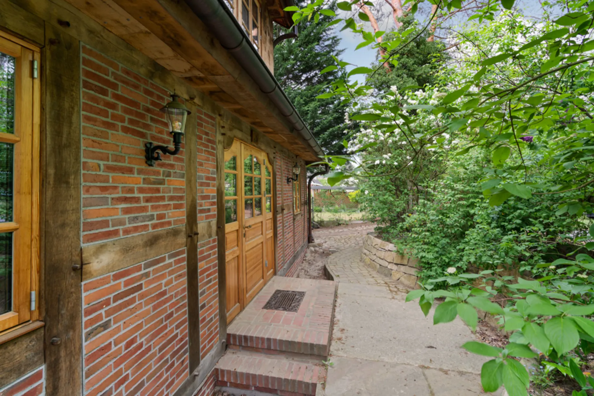 Backsteinfassade eines Hauses mit Holztür und Gartenweg, umgeben von grünen Pflanzen.