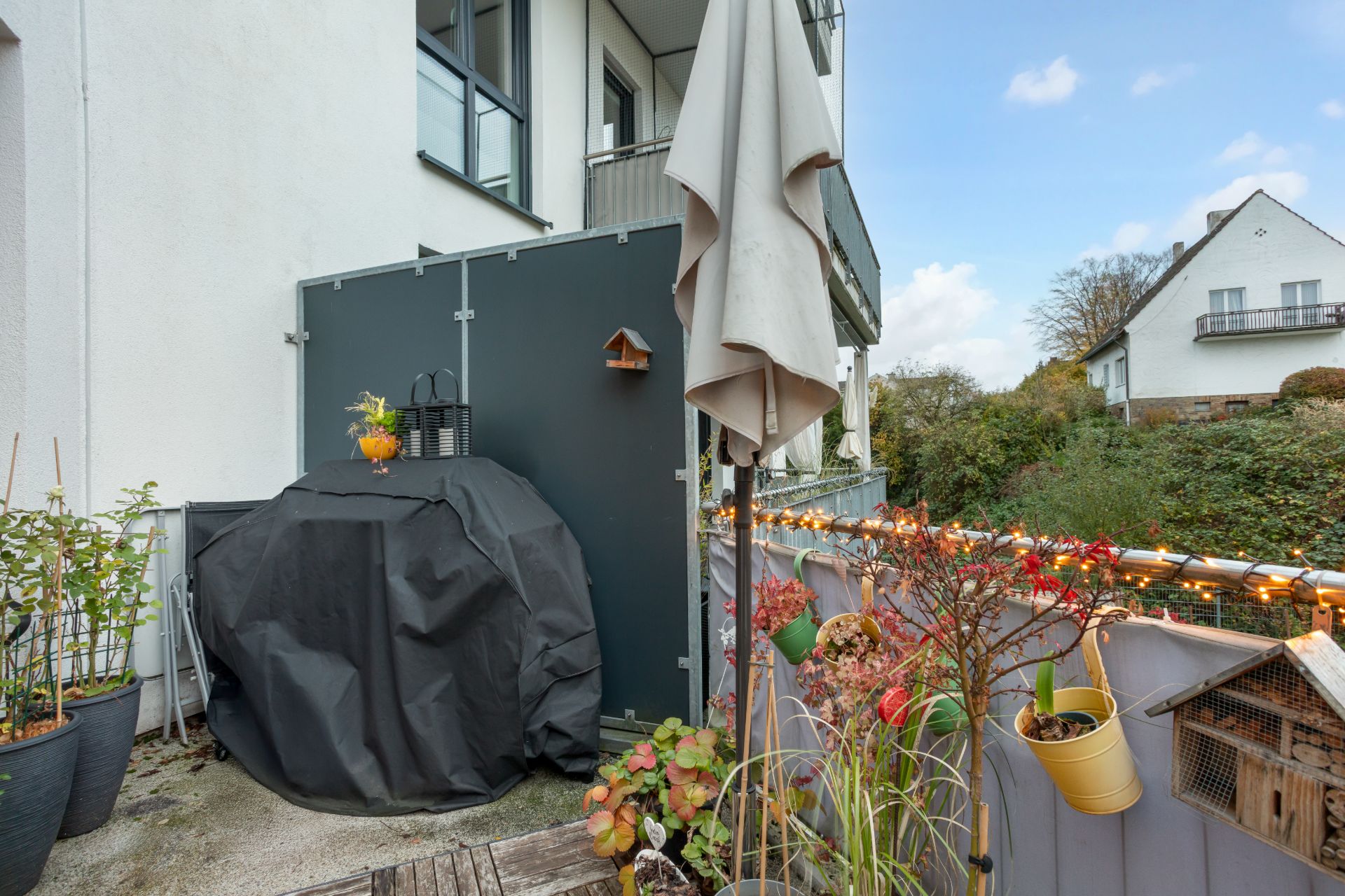Balkon mit Pflanzen, geschütztem Grill, geschlossener Markise und Lichterkette am Geländer.