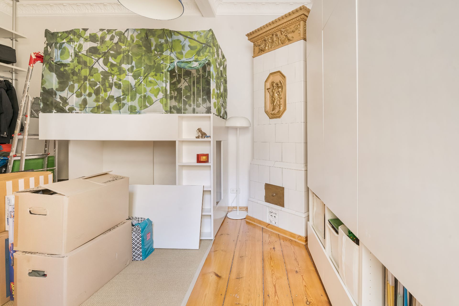 Zimmer mit Hochbett, Kartons, weißem Schrank und historischem Kachelofen.