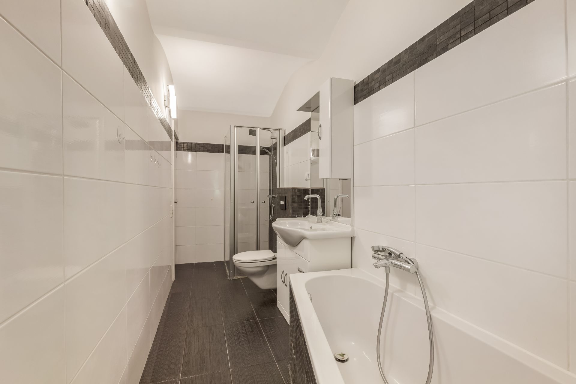 Modernes Badezimmer mit Badewanne, Dusche, WC und Waschbecken, weiße Fliesen und dunkler Boden.