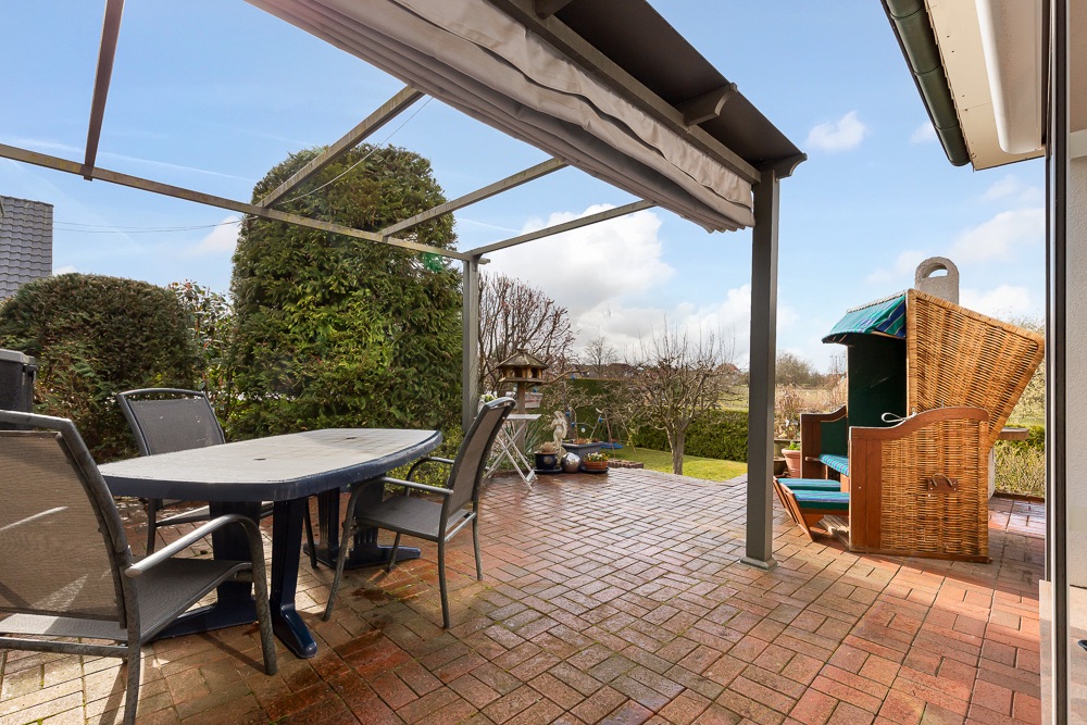 Überdachte Terrasse mit Tisch, Stühlen und Strandkorb, umgeben von Garten mit Büschen.