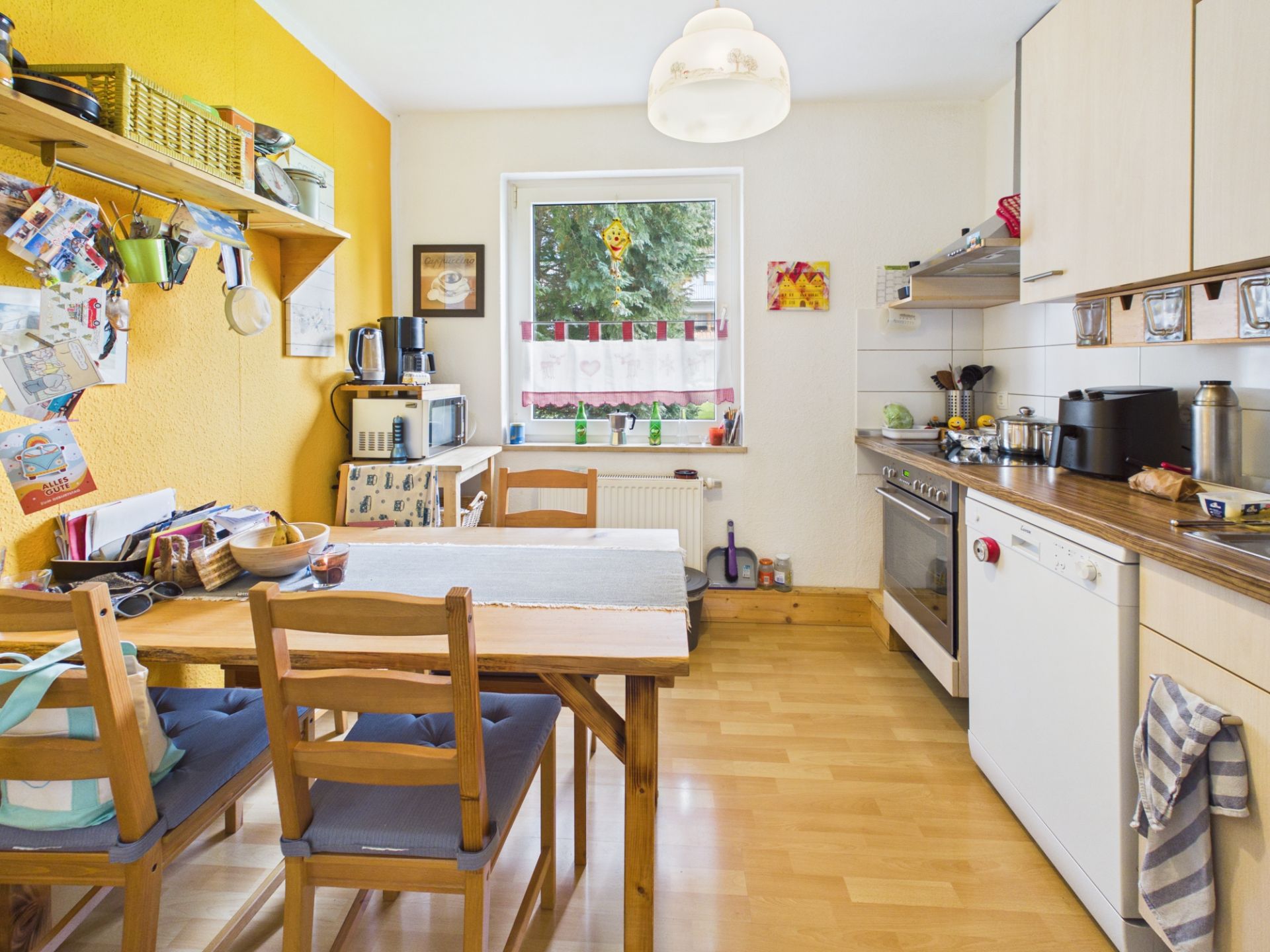 Helle Wohnküche mit Esstisch, vier Stühlen, gelber Wand und Fenster mit Vorhang.