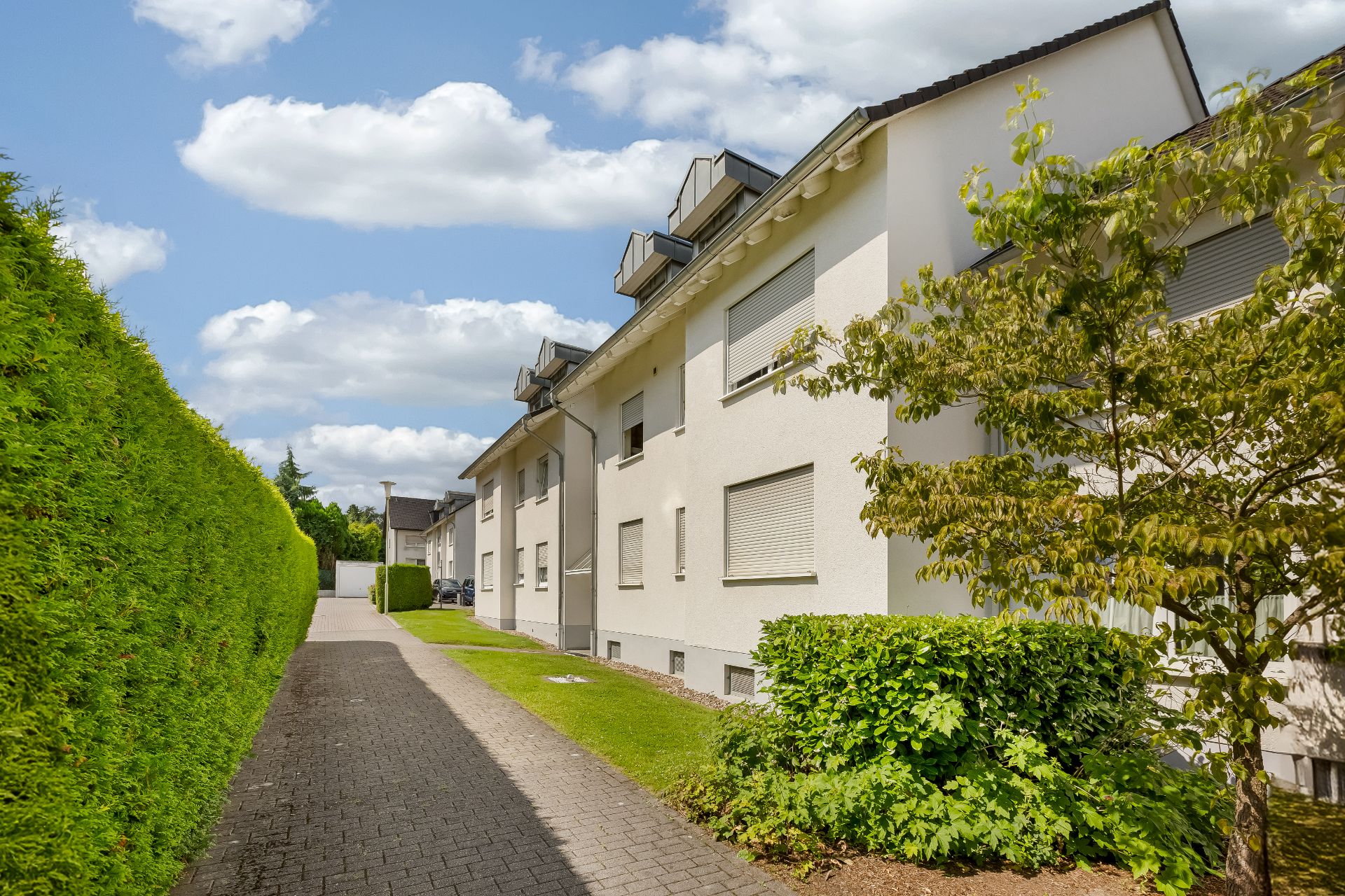 Gepflegter Weg neben einem weißen Wohnhaus mit Hecke und Bäumen bei Sonnenschein.