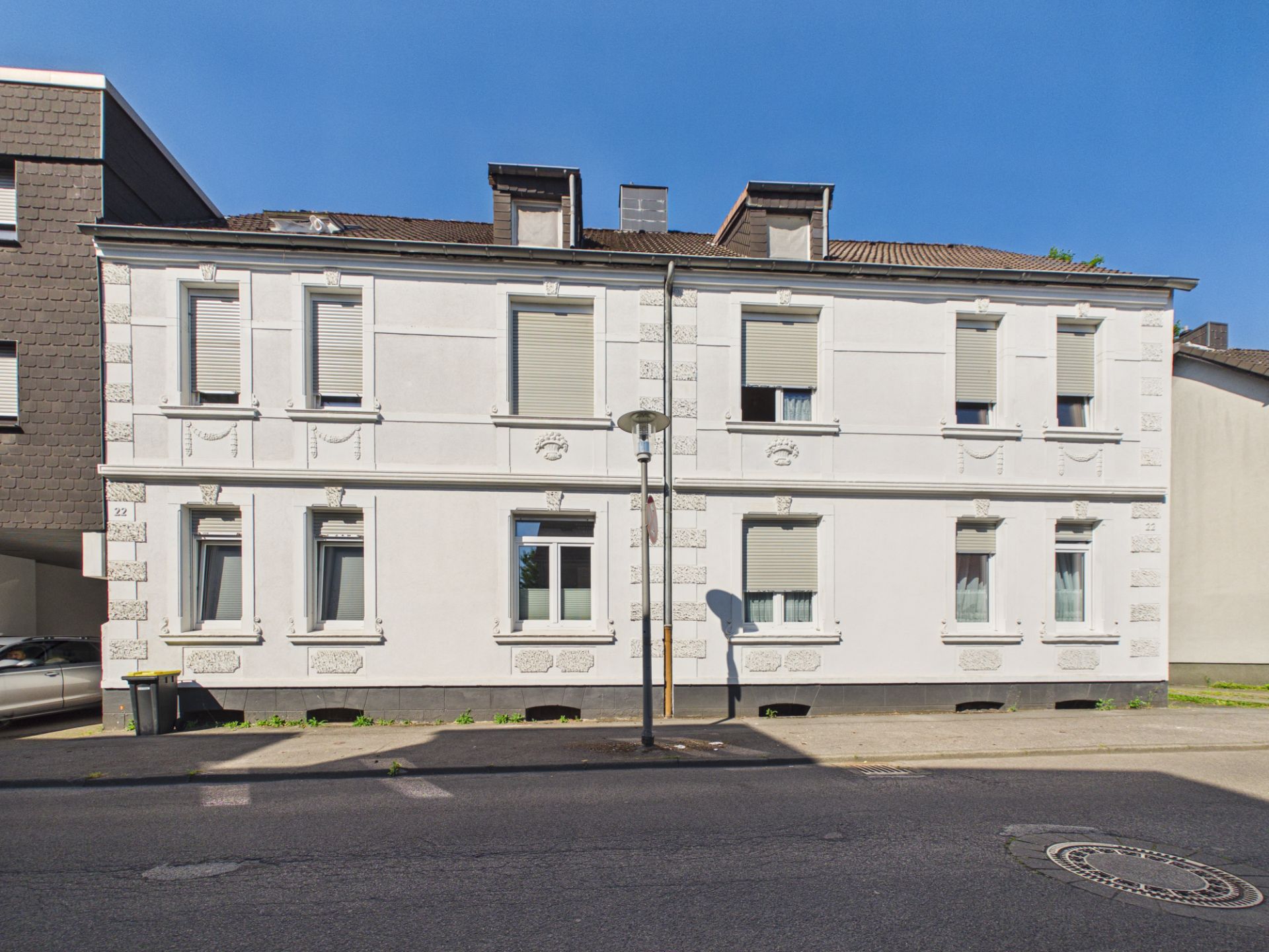 Zweistöckiges weißes Wohnhaus mit mehreren Fenstern und Rollläden an einer Straße.