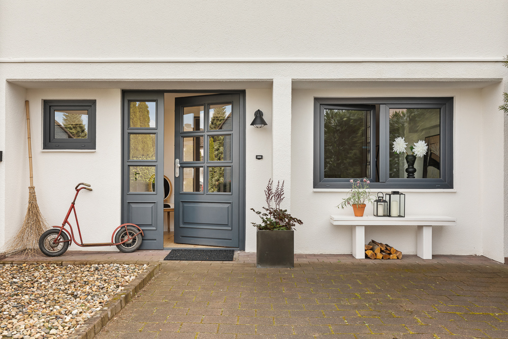 Eingangsbereich eines Hauses mit grauer Tür, Fenster, Roller, Bank, Pflanzen und Laternen.