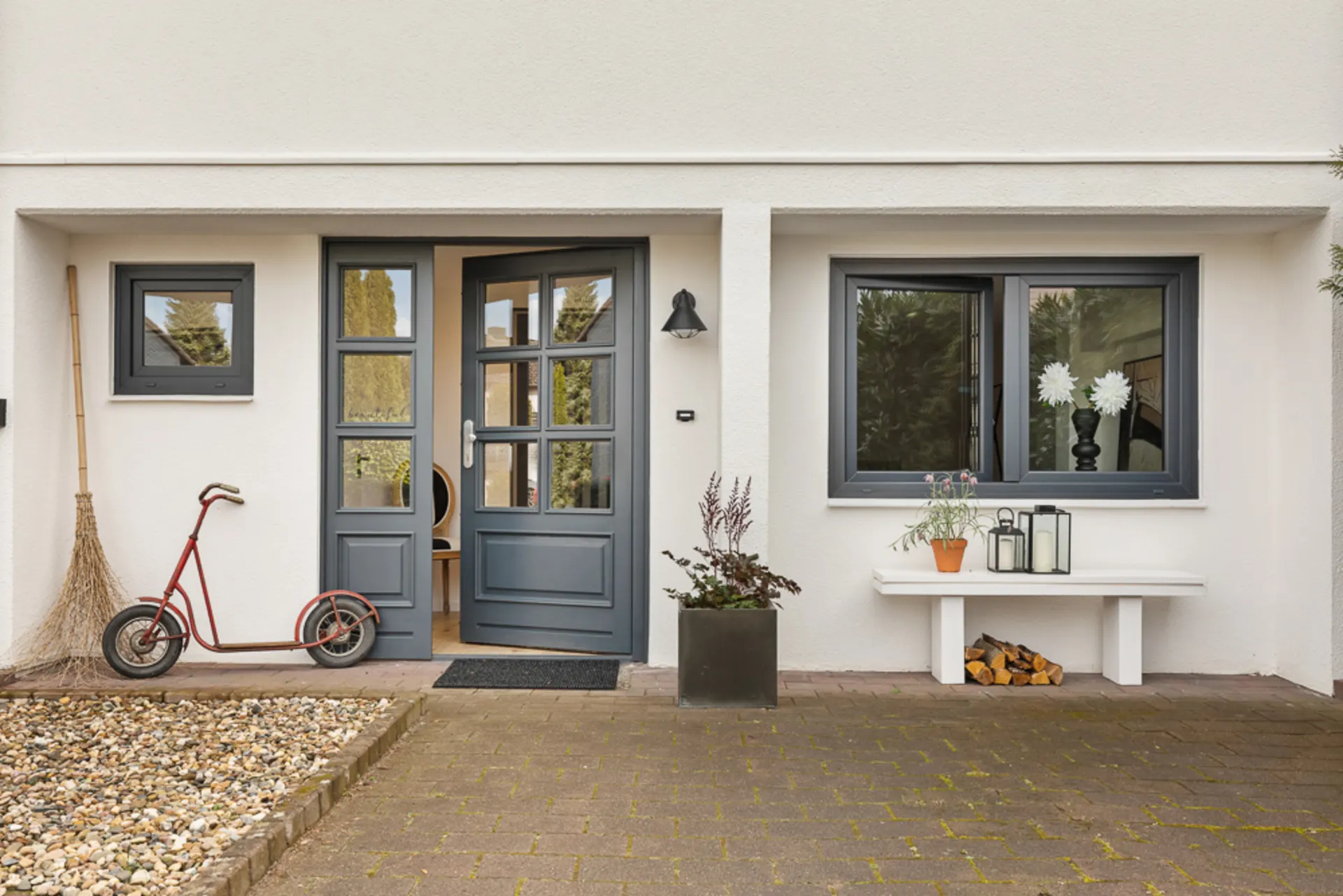 Eingangsbereich eines Hauses mit grauer Tür, Fenster, Roller, Bank, Pflanzen und Laternen.
