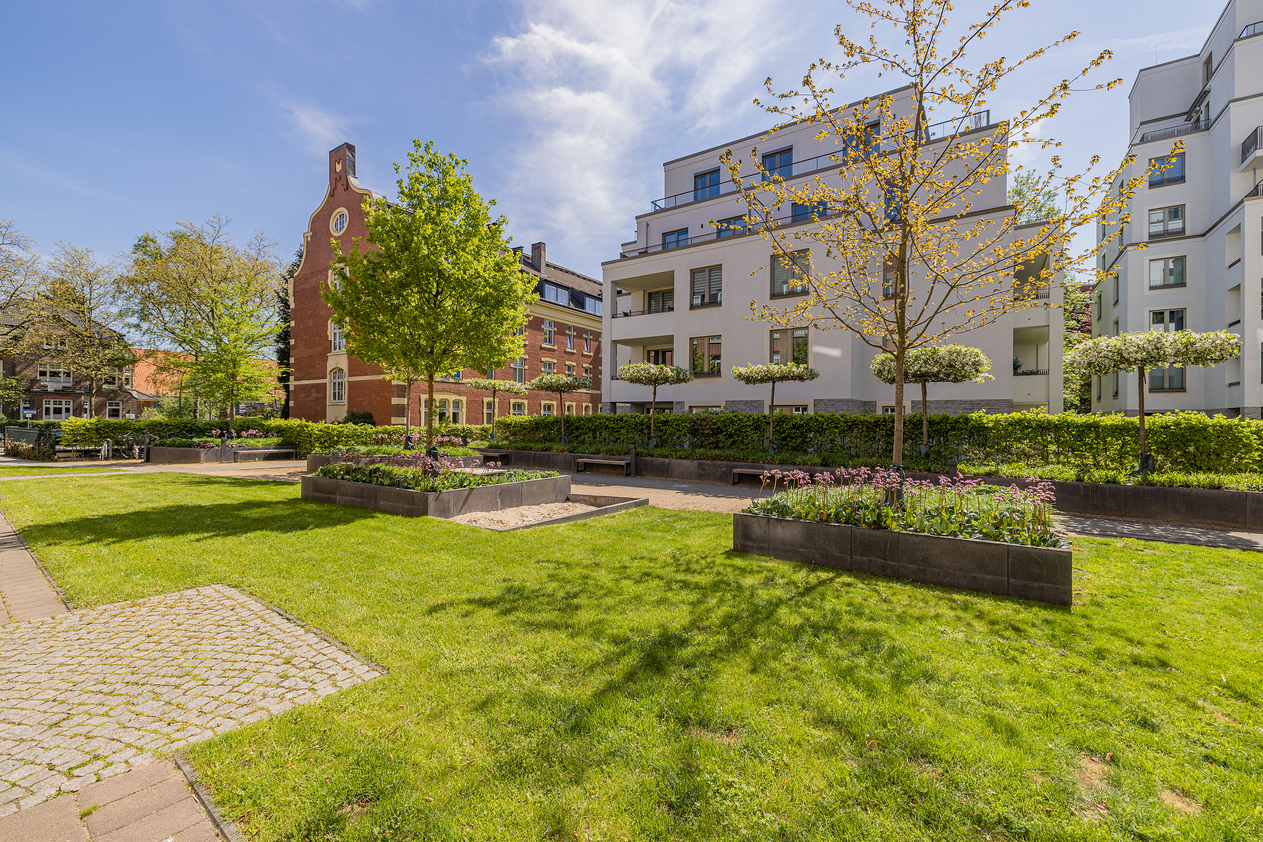 Grüne Wohnanlage mit modernen Gebäuden, Blumenbeeten und Bäumen bei sonnigem Wetter.