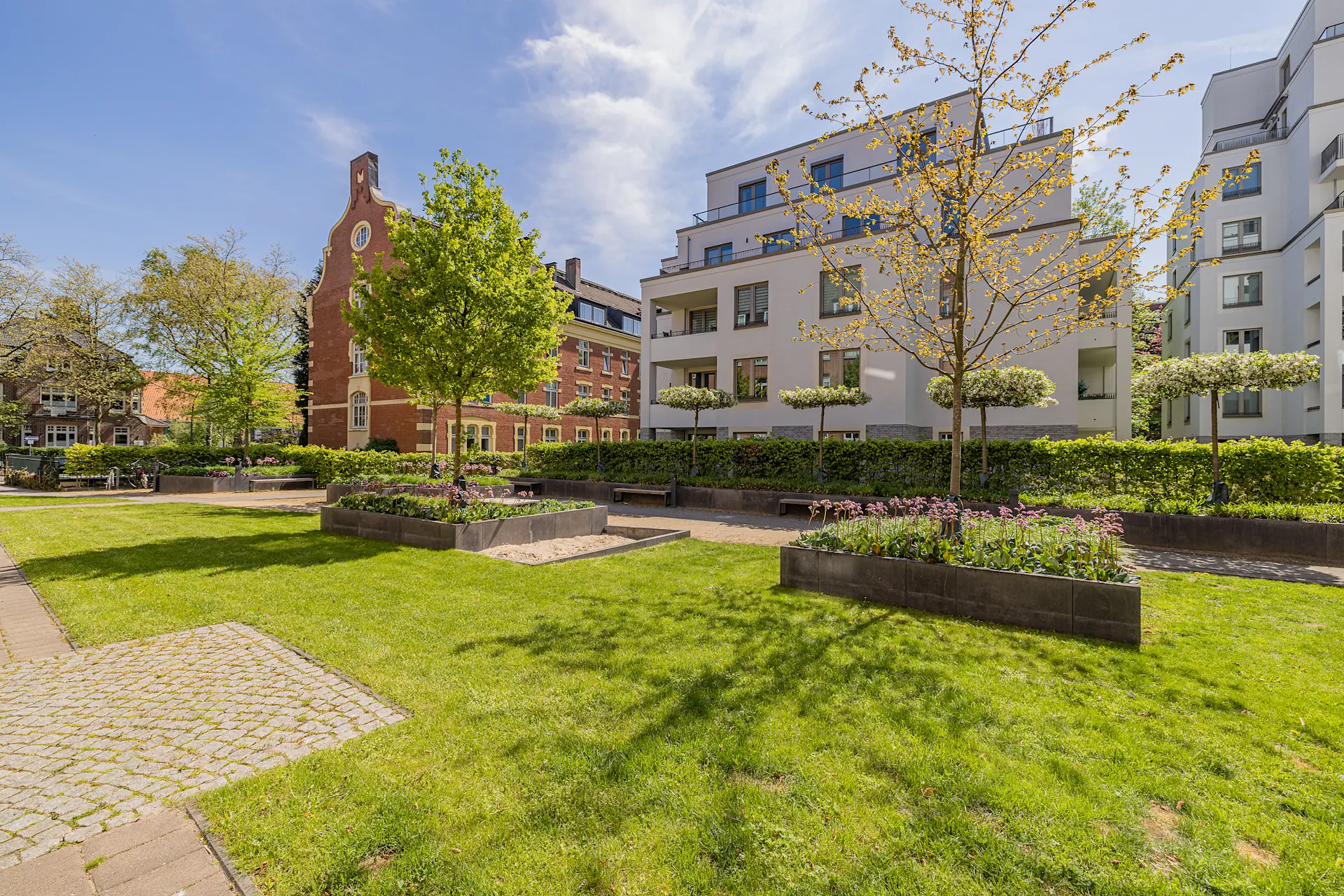 Grüne Wohnanlage mit modernen Gebäuden, Blumenbeeten und Bäumen bei sonnigem Wetter.