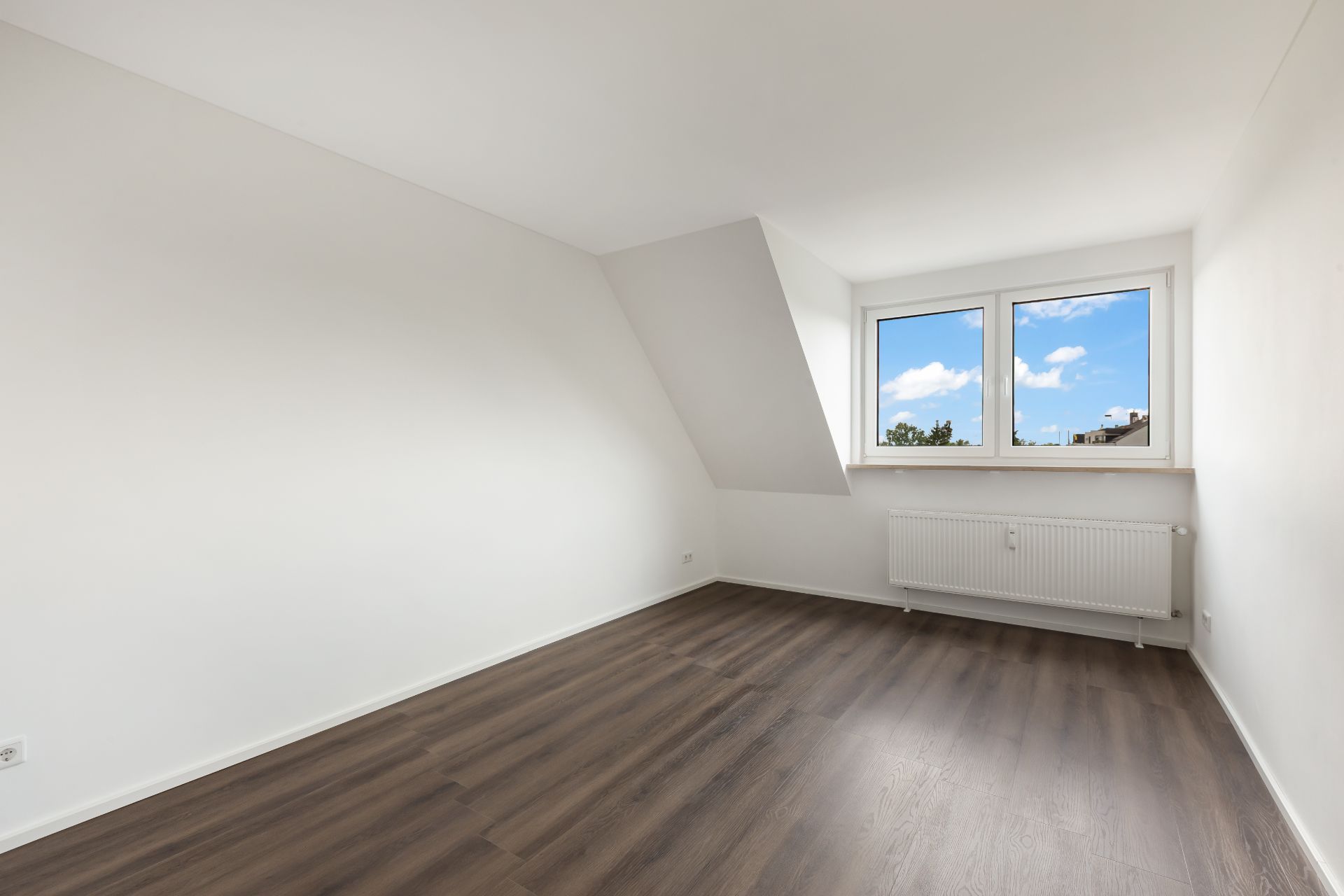 Leeres Zimmer mit weißen Wänden, dunklem Boden und Fenster mit Blick nach draußen.